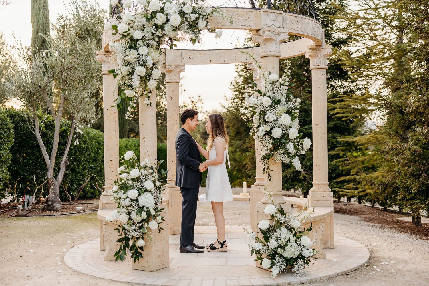 A romantic outdoor wedding ceremony setup with white floral arrangements adorning elegant stone columns at sunset.