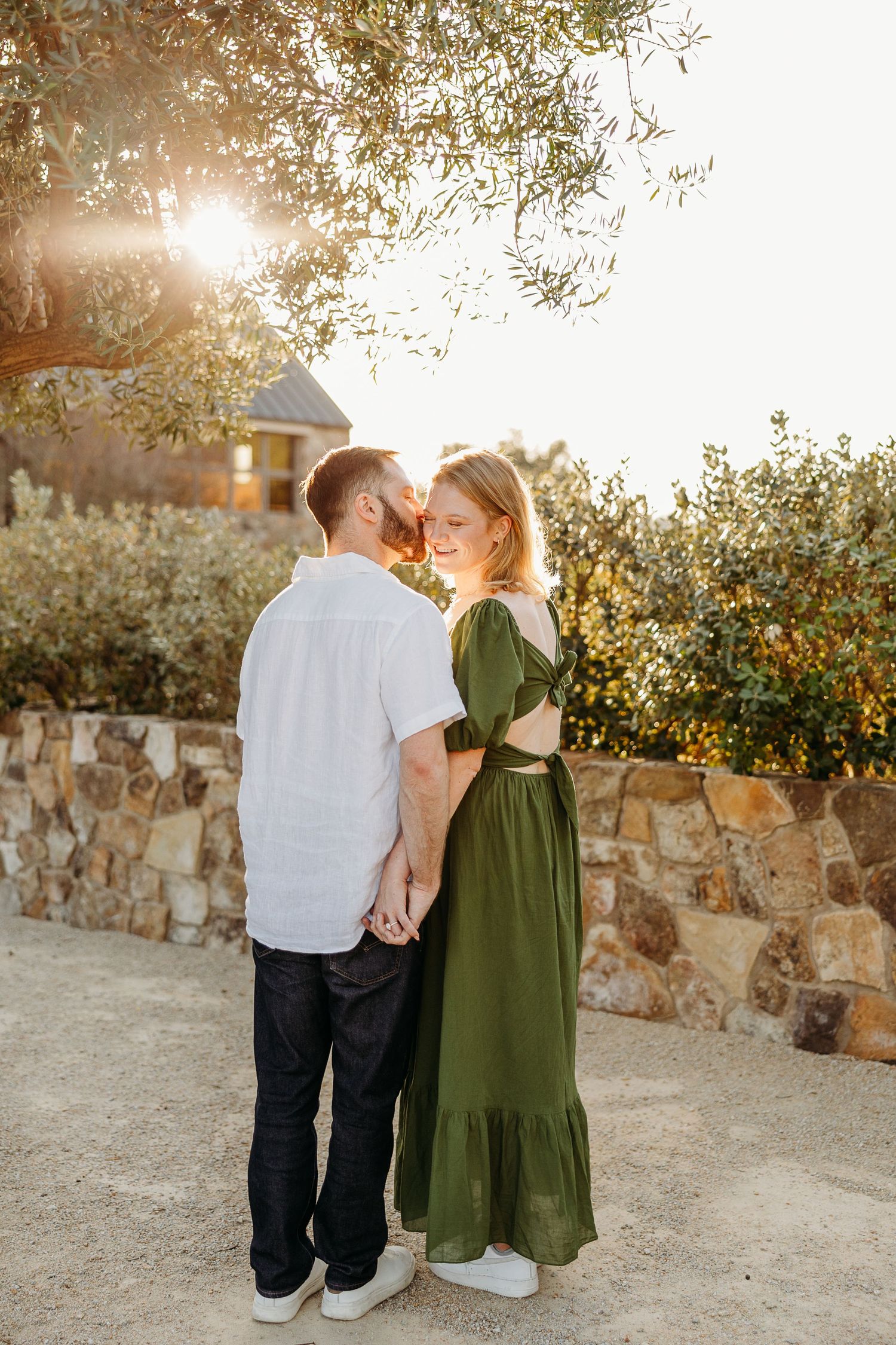 A couple in casual attire stands together against a rustic stone wall at sunset.