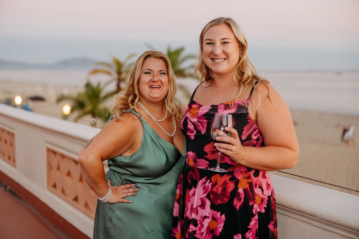 Two women in evening dresses smile while holding drinks on an oceanfront balcony at sunset.