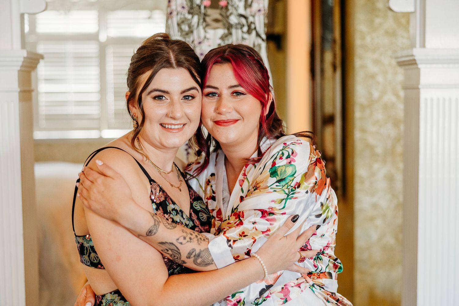 Two women in floral and embellished dresses embrace while posing together in a sunlit room.