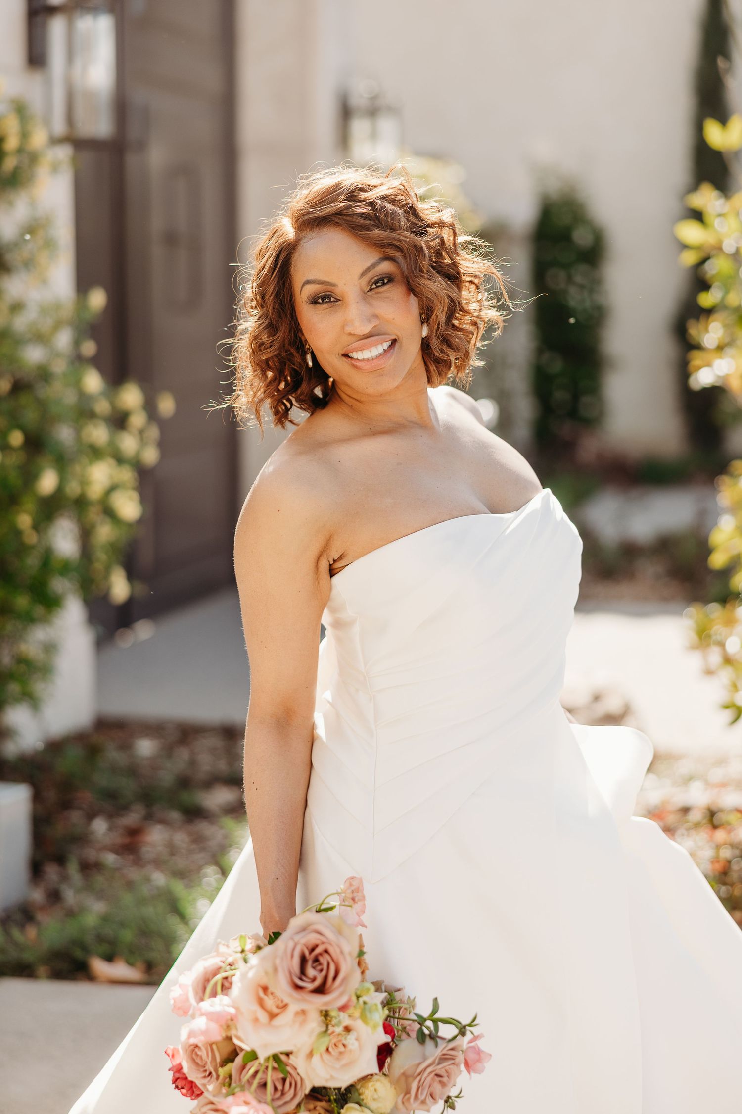 Bride in flowing white wedding gown holds pink and blush rose bouquet in sunlit garden setting.