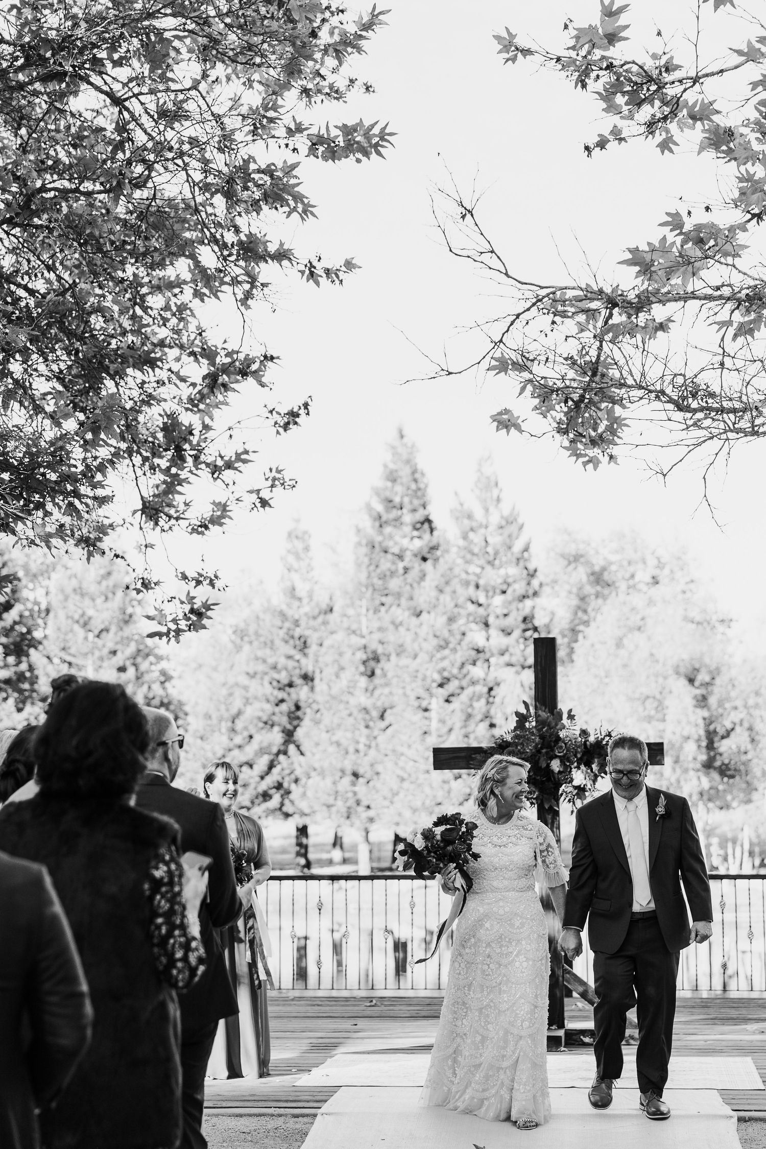 A black and white wedding ceremony photo with a cross backdrop and pine trees in the background.