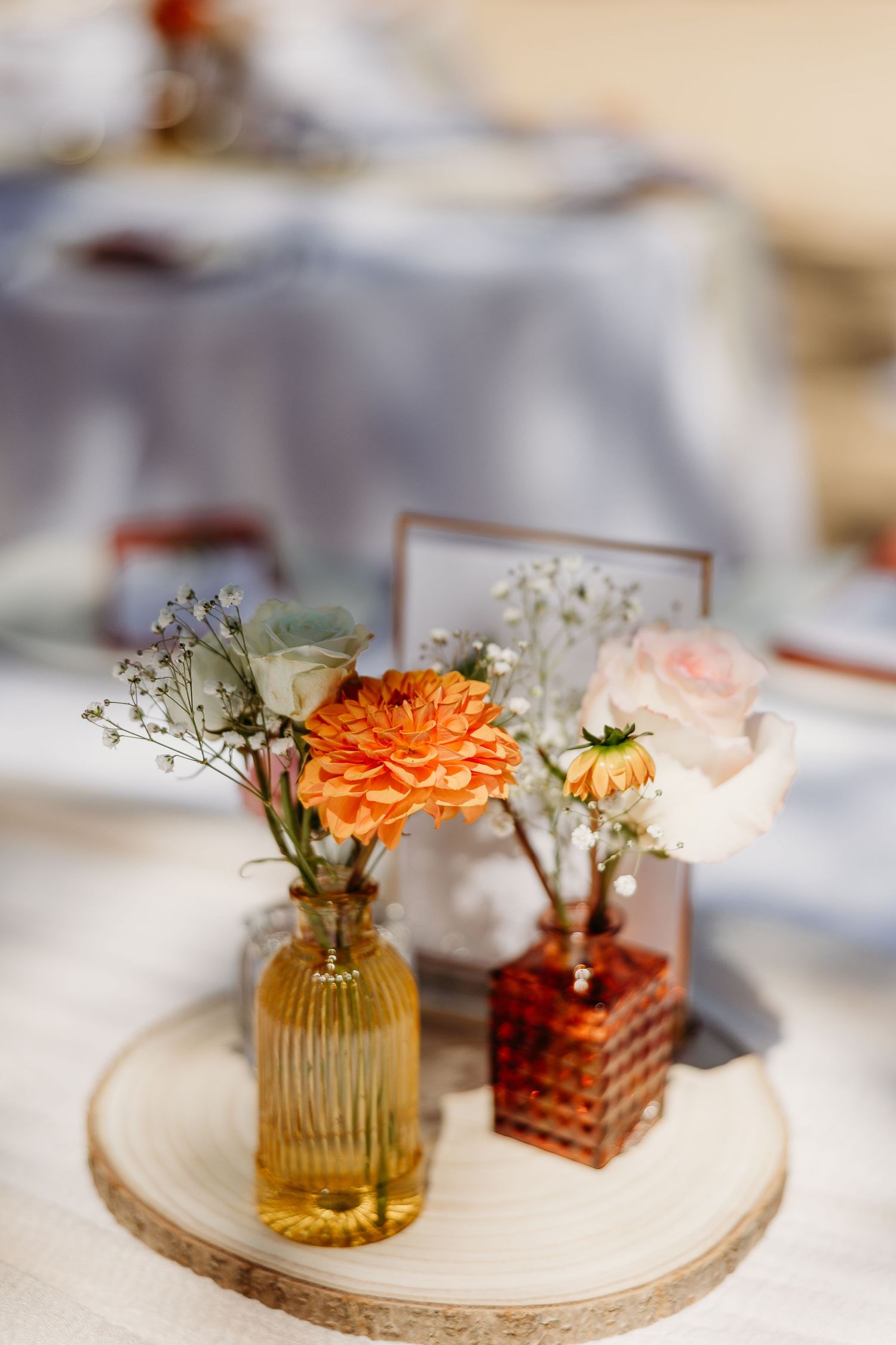 Delicate floral centerpiece with orange dahlia in amber glass vase on wooden slice.