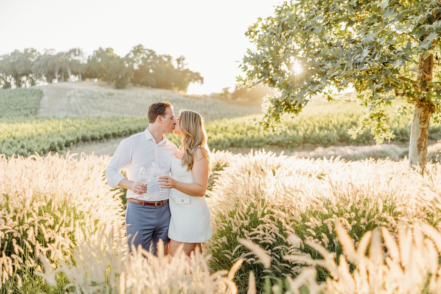 Romantic couple embraces in golden wheat field at sunset with rolling hills in background.