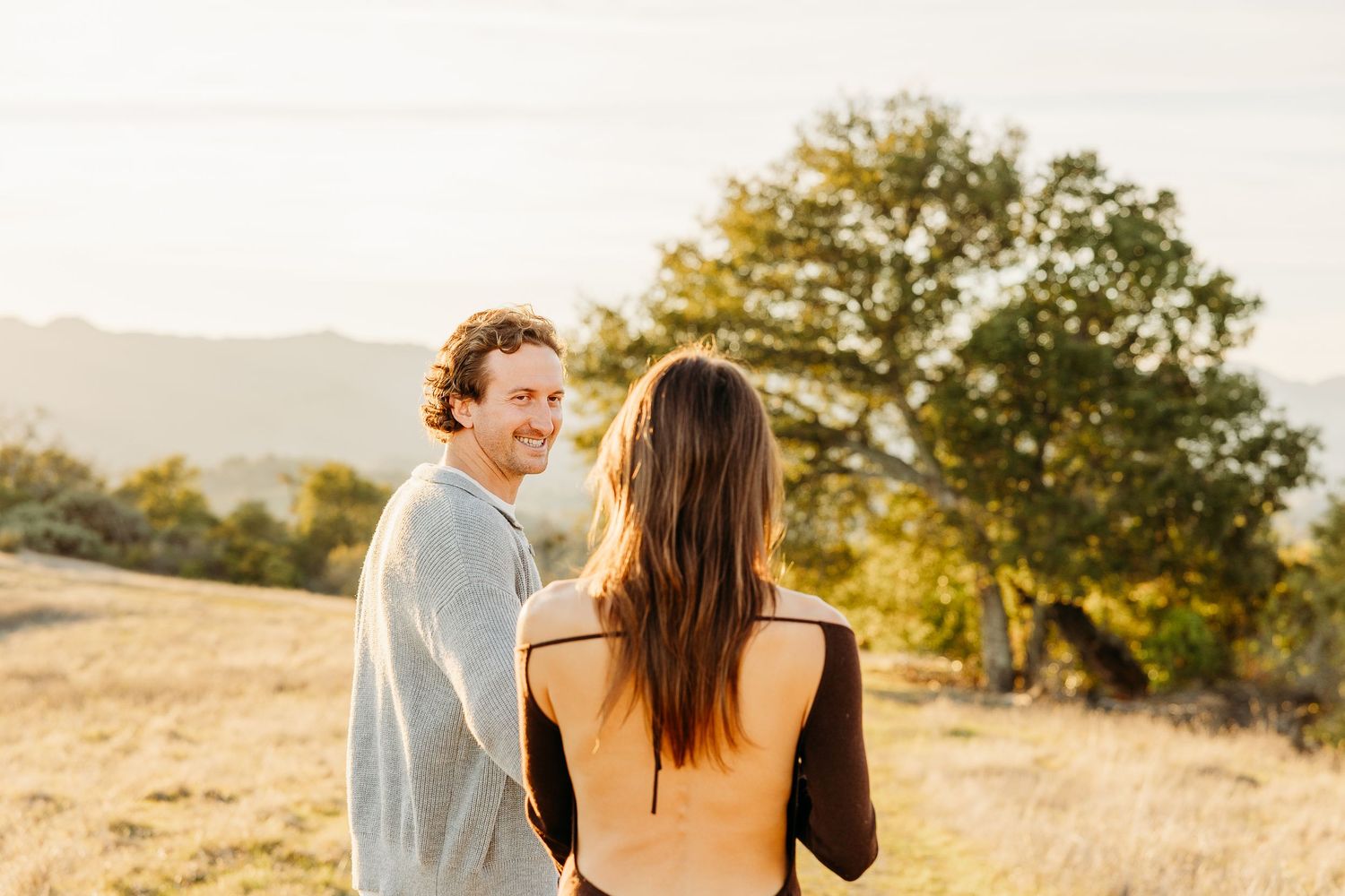 Two people share an intimate moment at sunset with rolling hills in the background.
