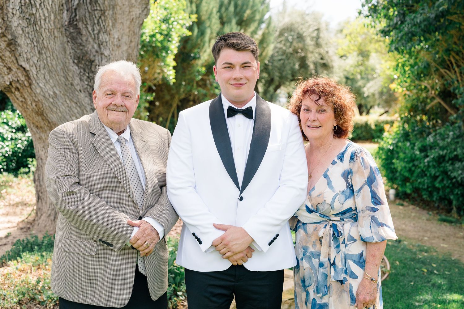 Groom in white tuxedo posing with parents outdoors under a tree.