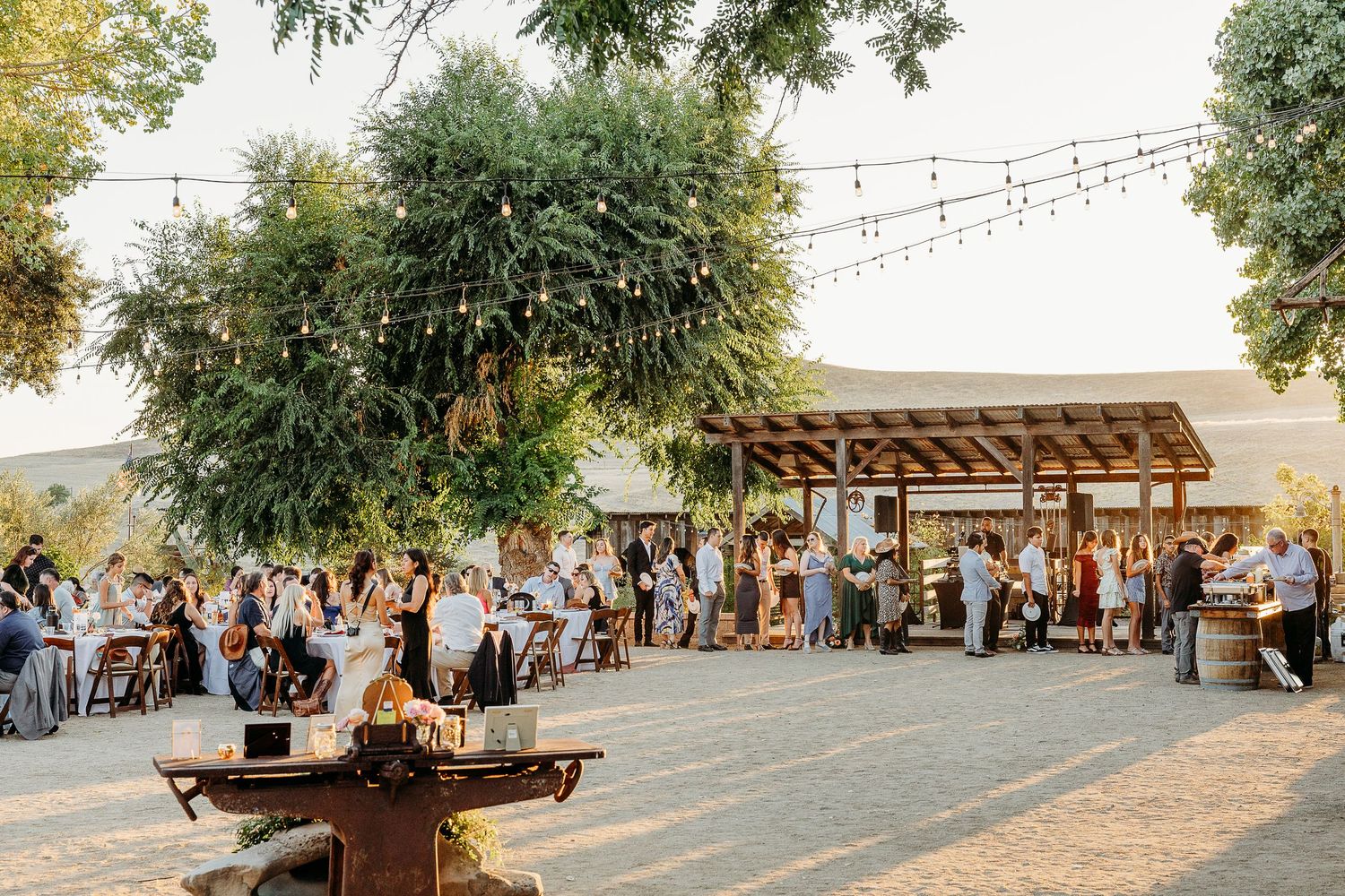 A rustic outdoor wedding reception venue with string lights and wooden tables set under tall trees.