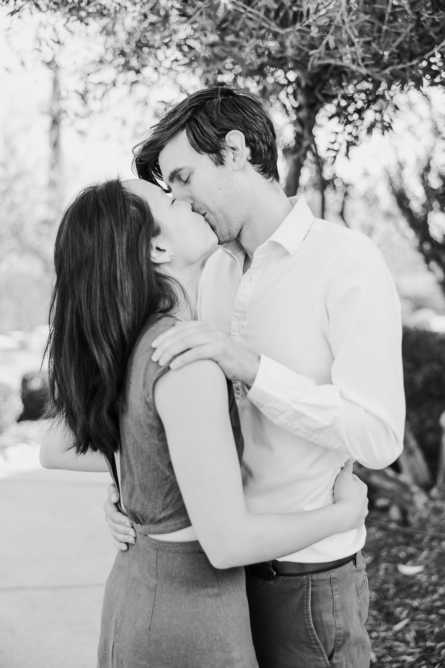 A romantic black and white portrait shows a tender embrace and kiss between a couple outdoors among trees.