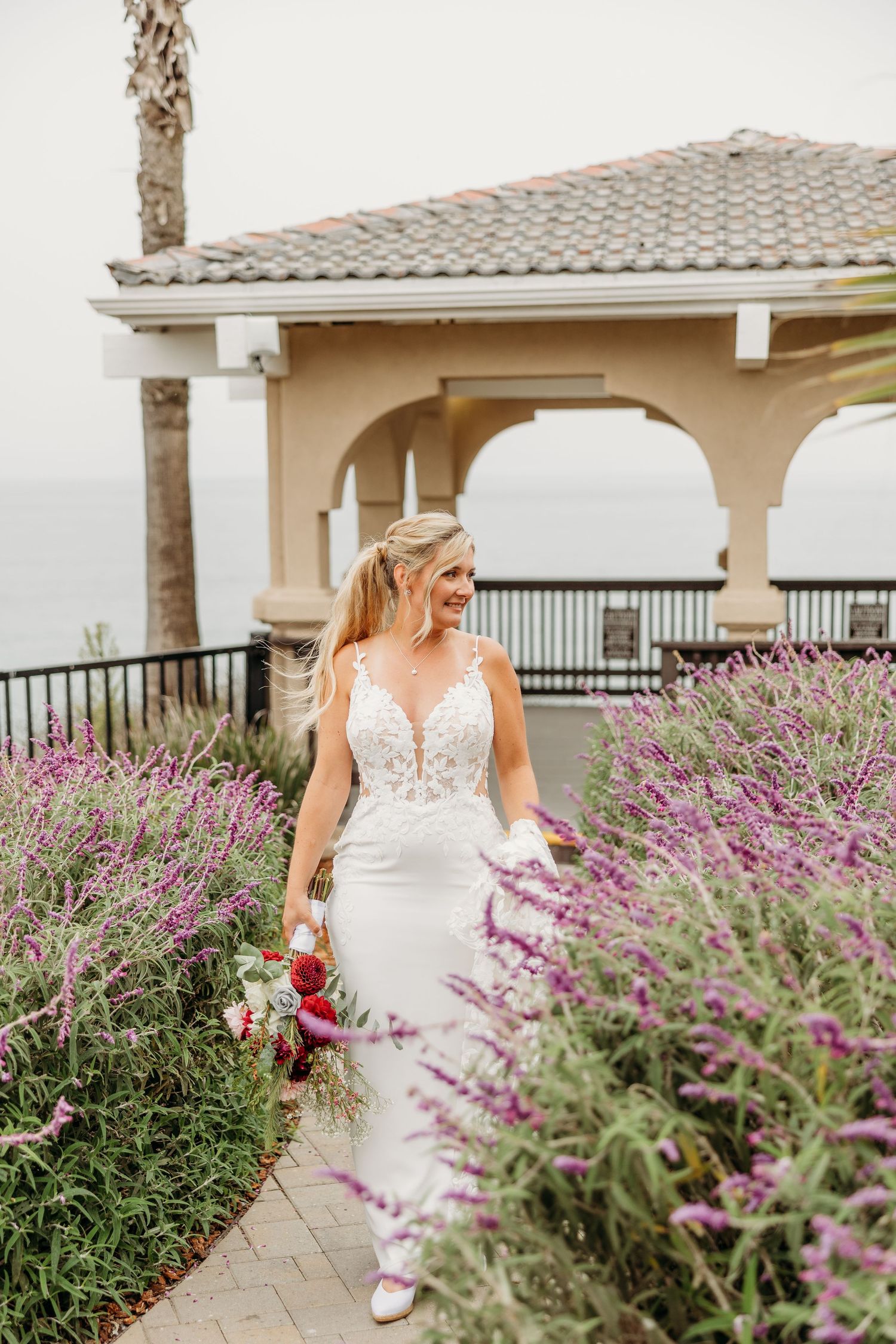 Elegant bridal portrait along garden path with blooming purple flowers and coastal gazebo.