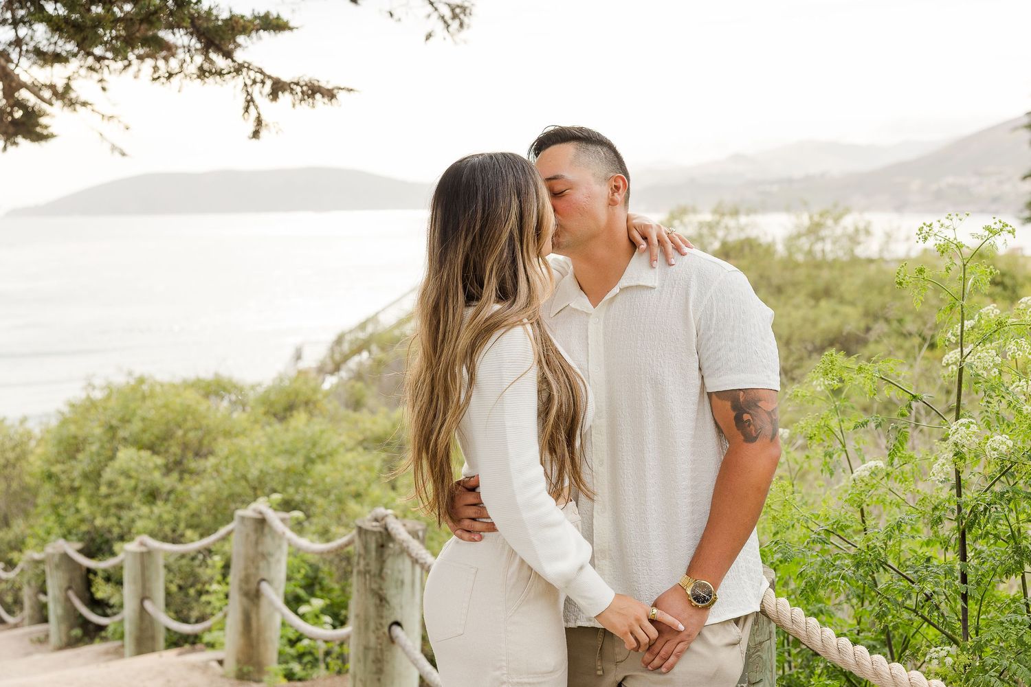 A couple in white outfits shares a romantic moment on a coastal cliffside with ocean views in the background.