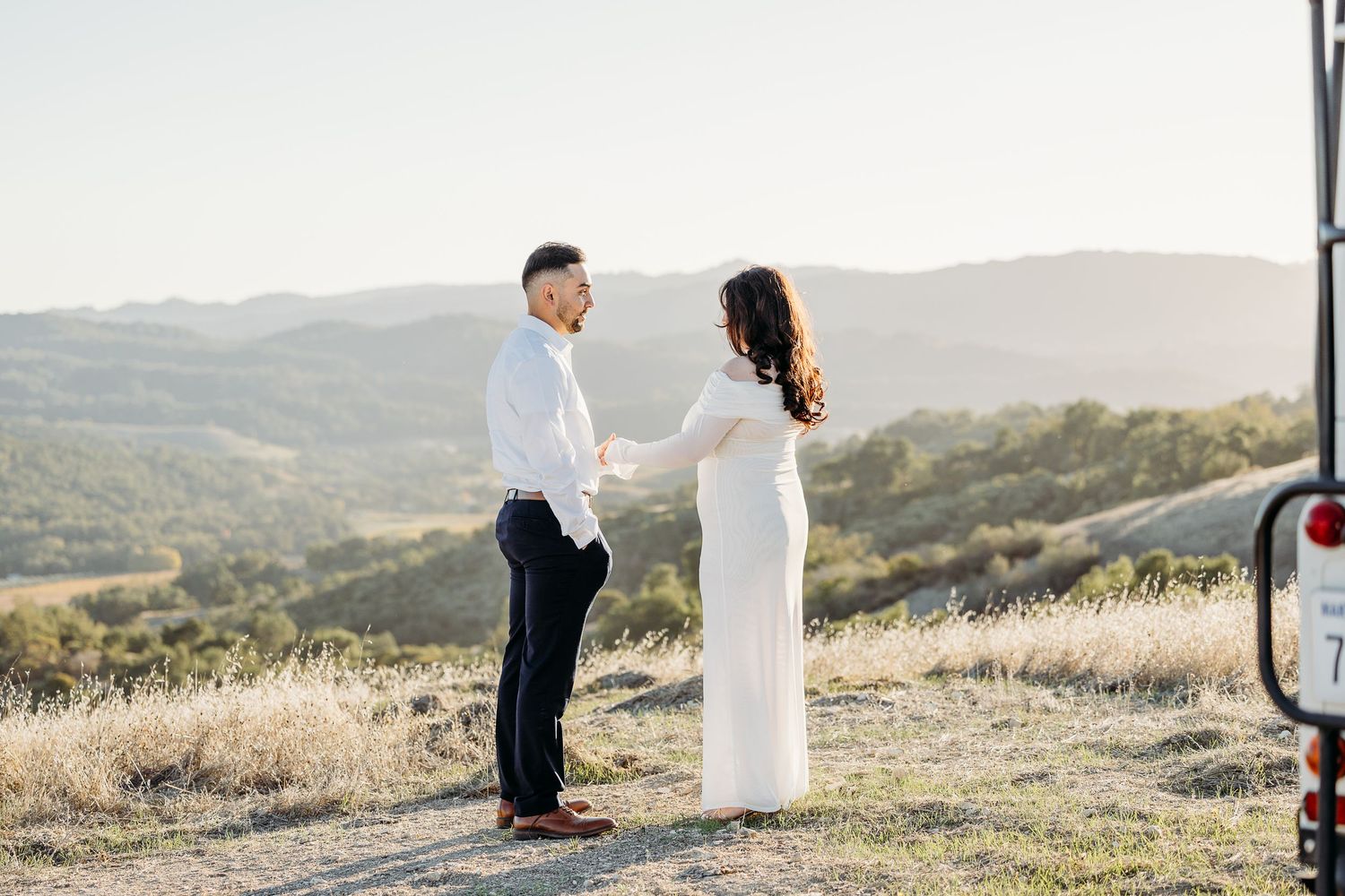 A couple in wedding attire stands together on a scenic hilltop overlooking rolling mountains at sunset.