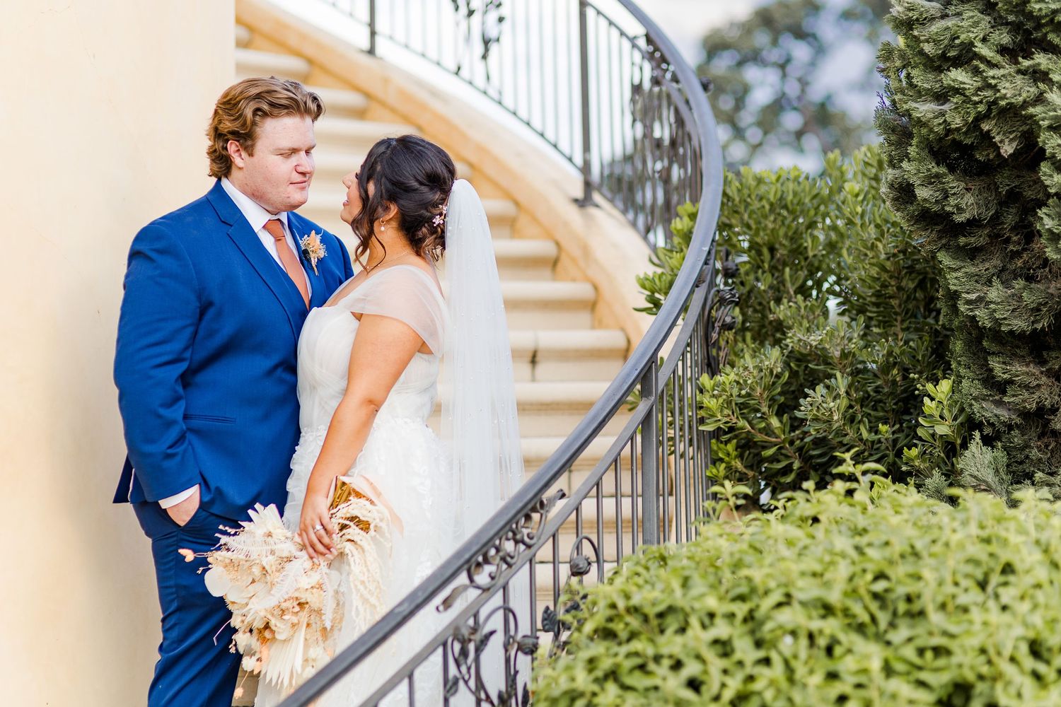 Newlyweds pose on elegant curved stairs with a dried flower bouquet.