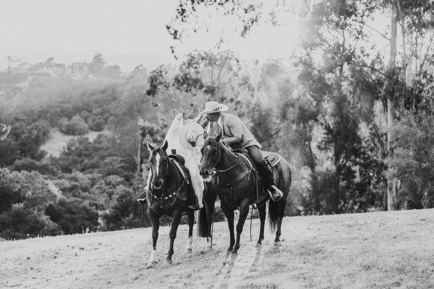 Two riders on horseback traverse a misty hillside path in black and white.