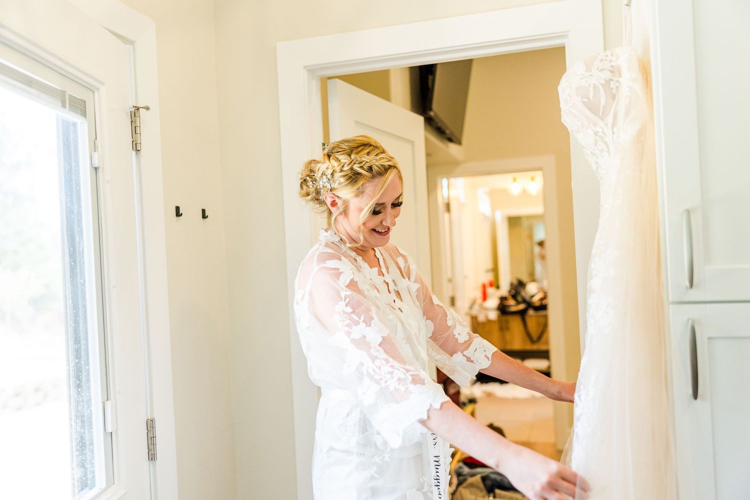 A person in white lace examines a wedding dress hanging in a bright hallway.