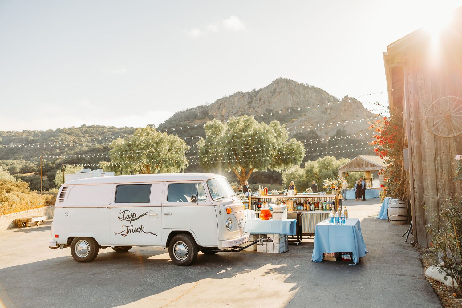 A vintage white VW van parked in front of a mountain with an outdoor dining setup at sunset.