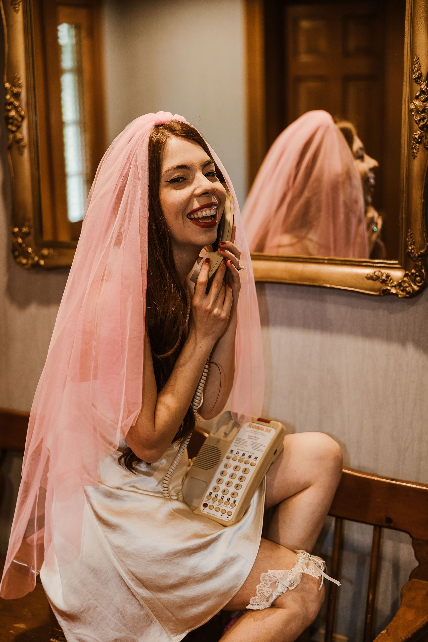 A bride in a pink veil laughs while holding a vintage telephone in front of an ornate gold mirror.