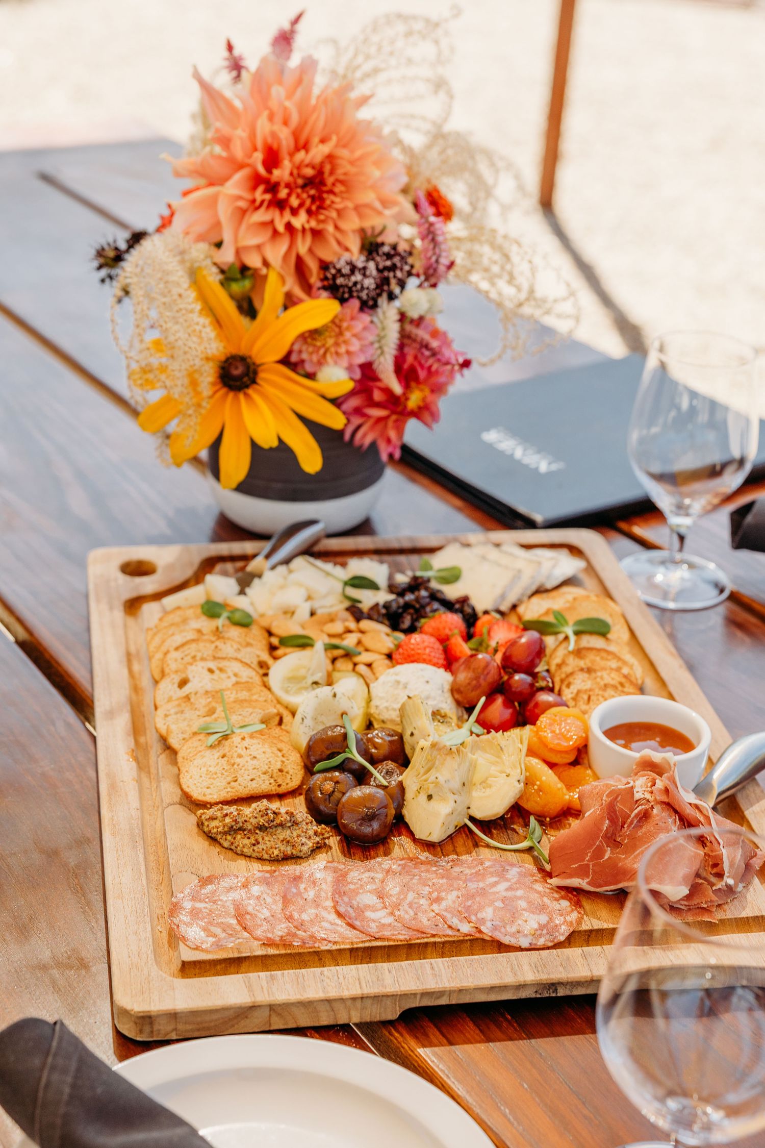 Wooden charcuterie board with meats, cheeses, and colorful flowers in vases arranged on rustic table setting.