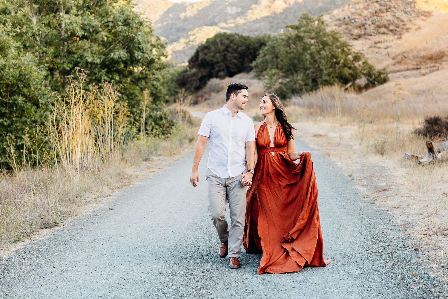 Couple strolls hand in hand on rural path surrounded by golden hills.