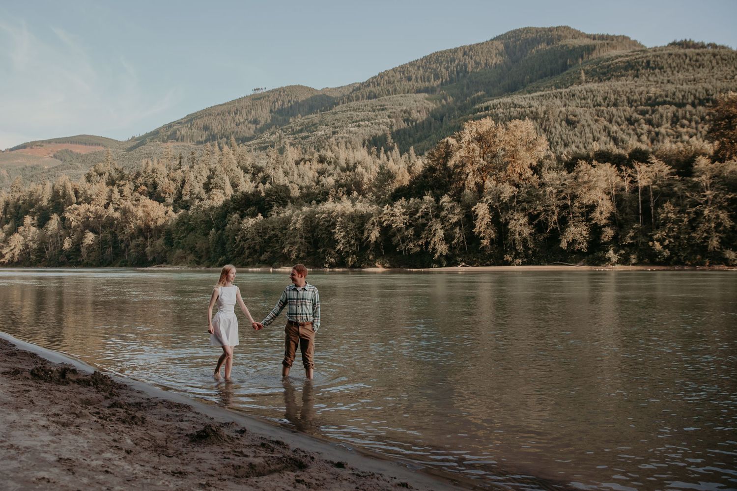 A couple walks along the rocky shoreline of a scenic mountain lake surrounded by evergreen trees and rugged peaks.