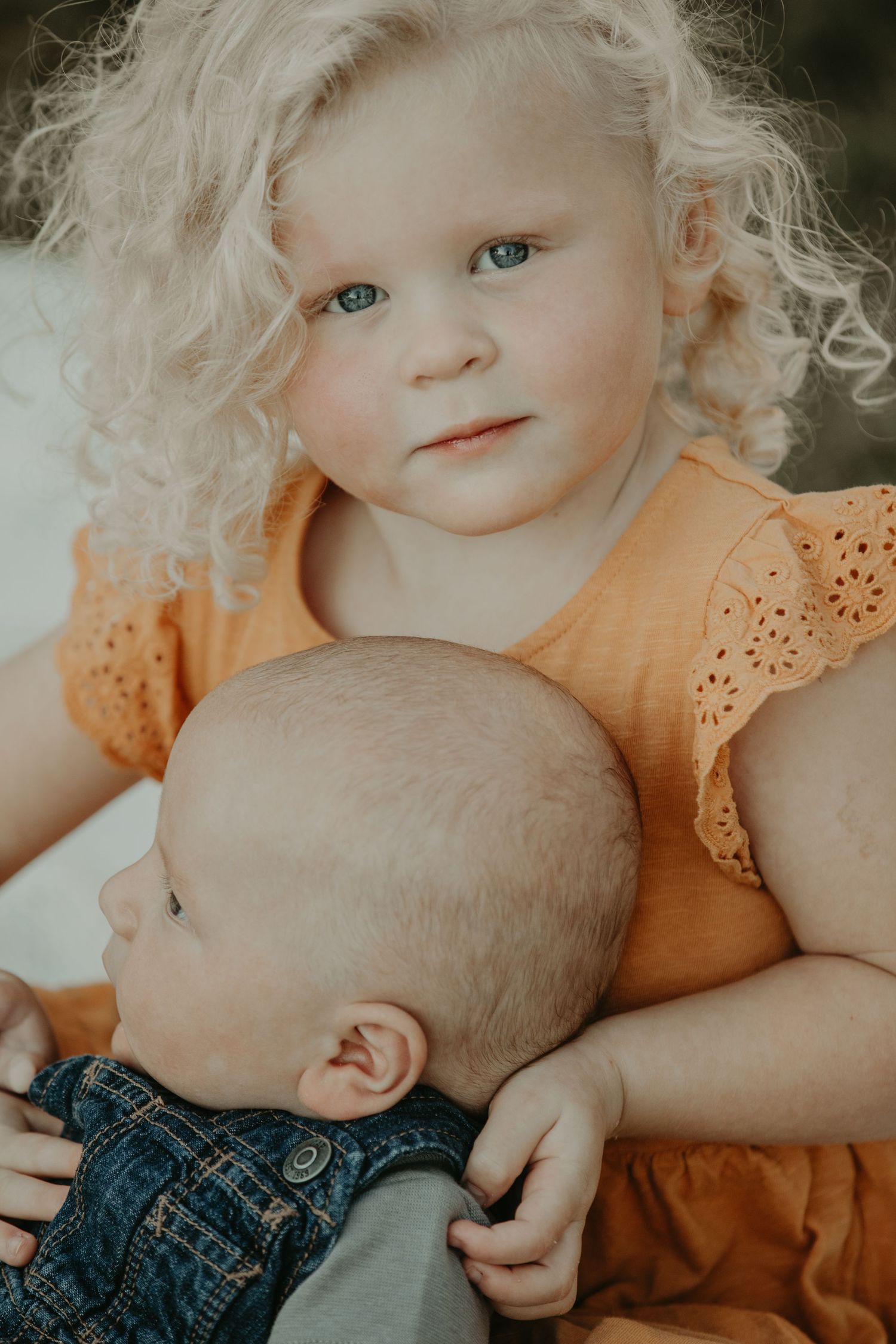 Two children, one with light hair in an orange top, lay close together tenderly on a blanket outdoors.