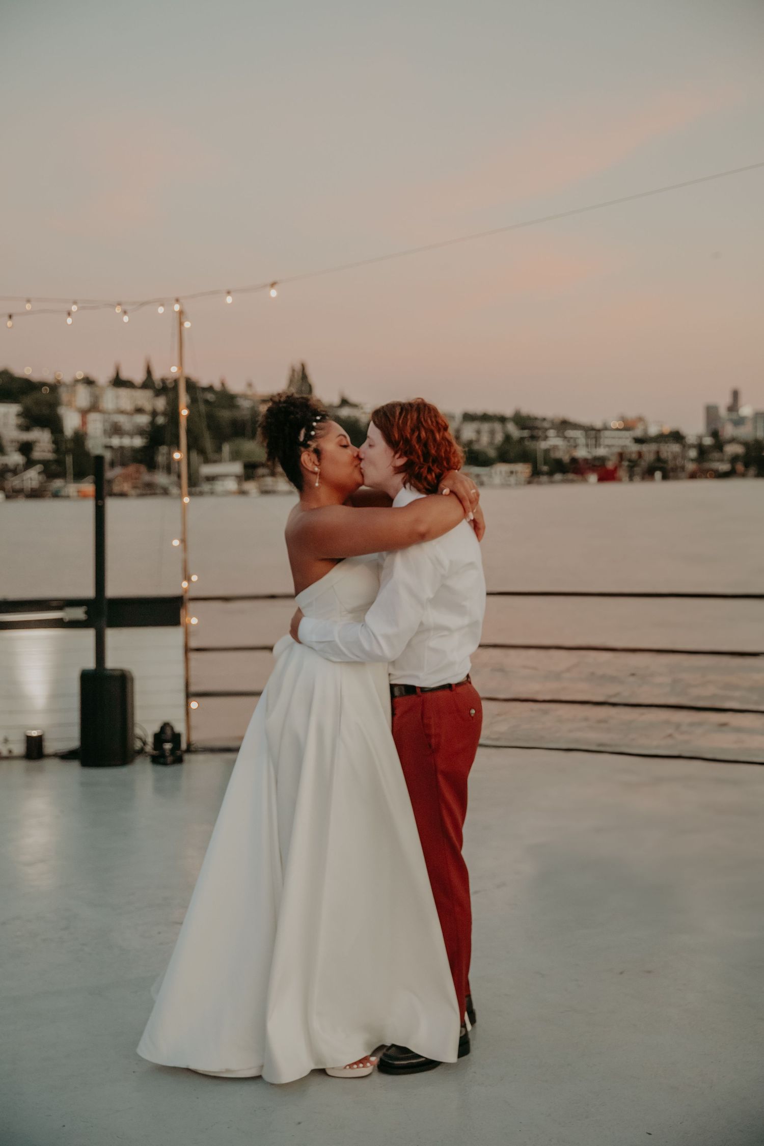 A romantic couple shares their first dance at sunset on a waterfront deck with string lights overhead and city views.