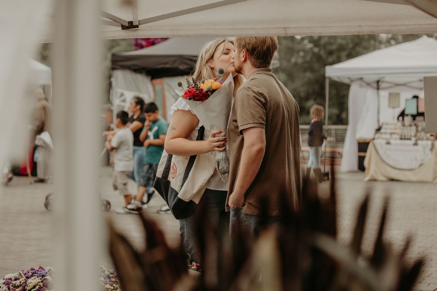 A couple embraces while dancing outdoors at a summer event with white tents and other attendees visible in background.