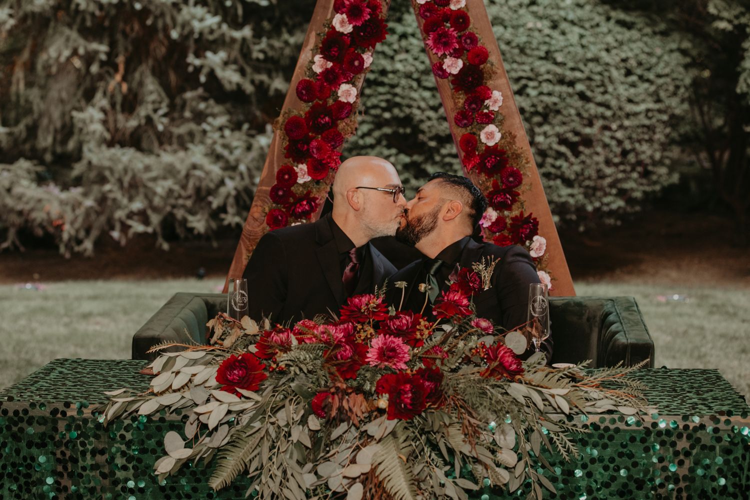 A romantic outdoor wedding ceremony setup with a floral arch and red roses against a moody forest backdrop.