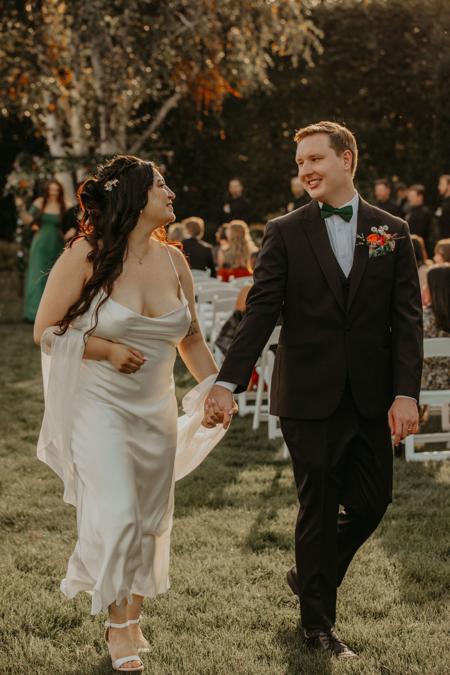 Bride and groom walk hand-in-hand on grass after outdoor wedding ceremony, smiling joyfully in elegant attire.