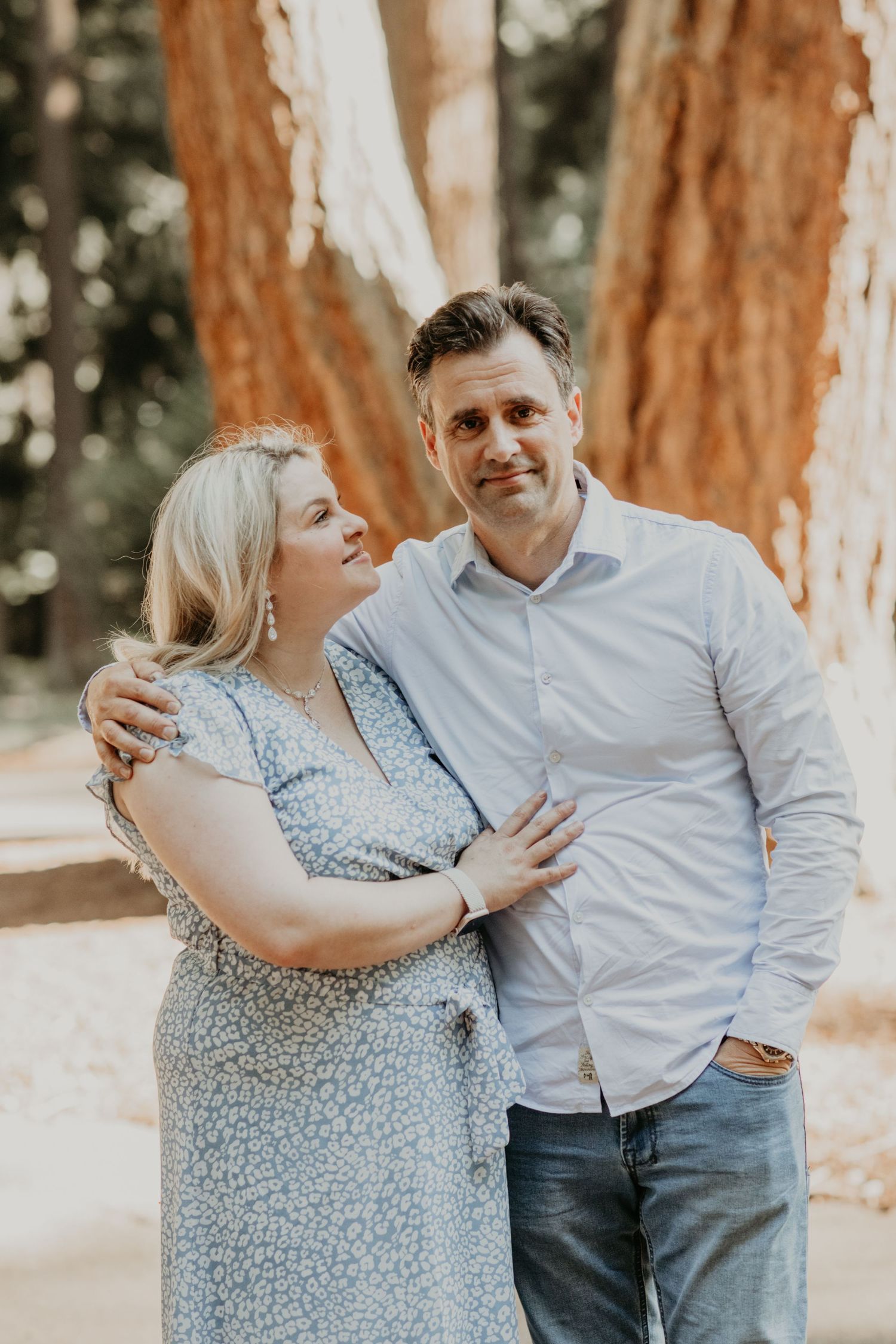 A romantic couple embraces outdoors in front of tall redwood trees on a sunny afternoon.