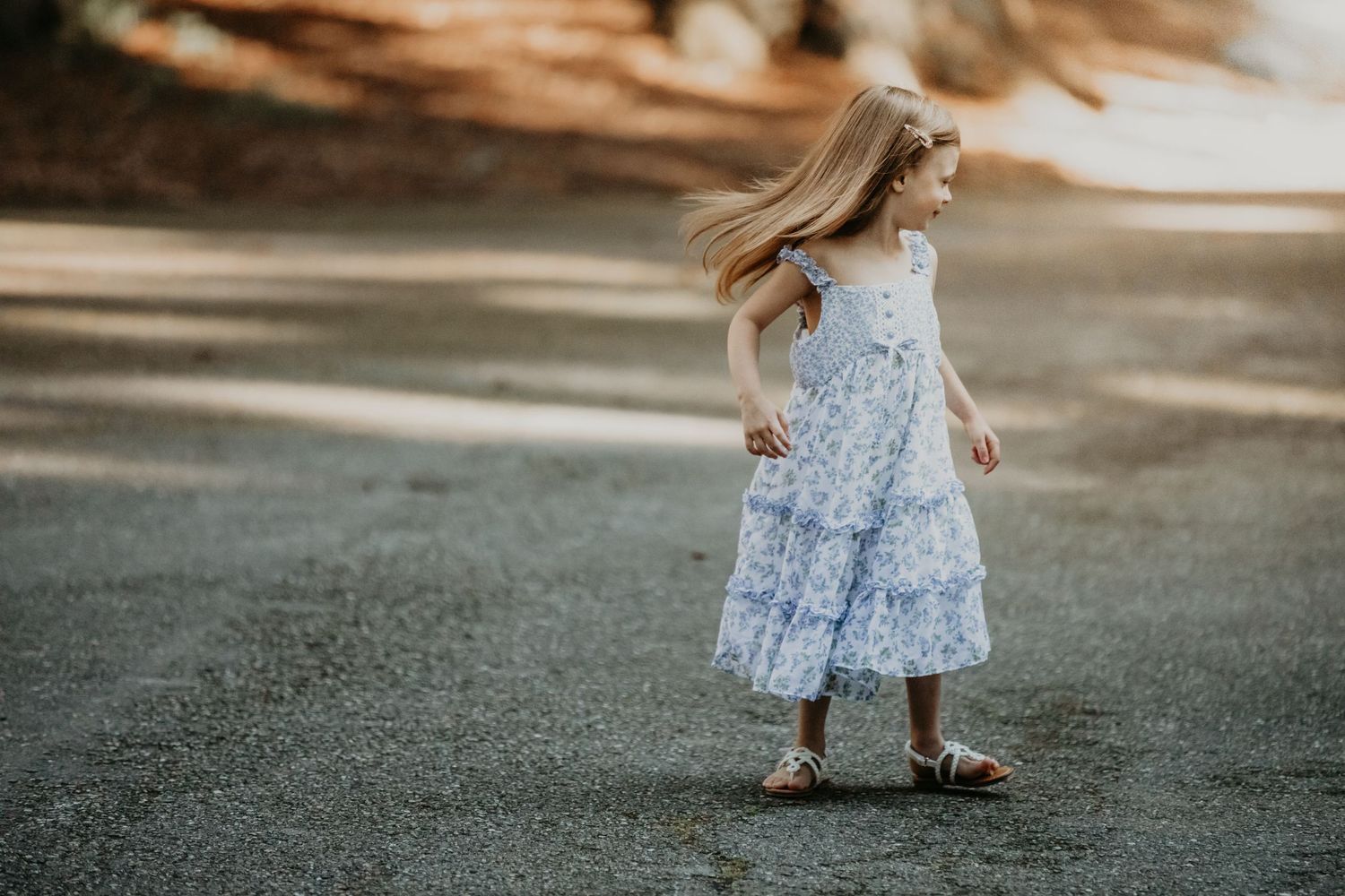 Young girl in a light blue floral dress twirls outdoors on asphalt during golden hour lighting.