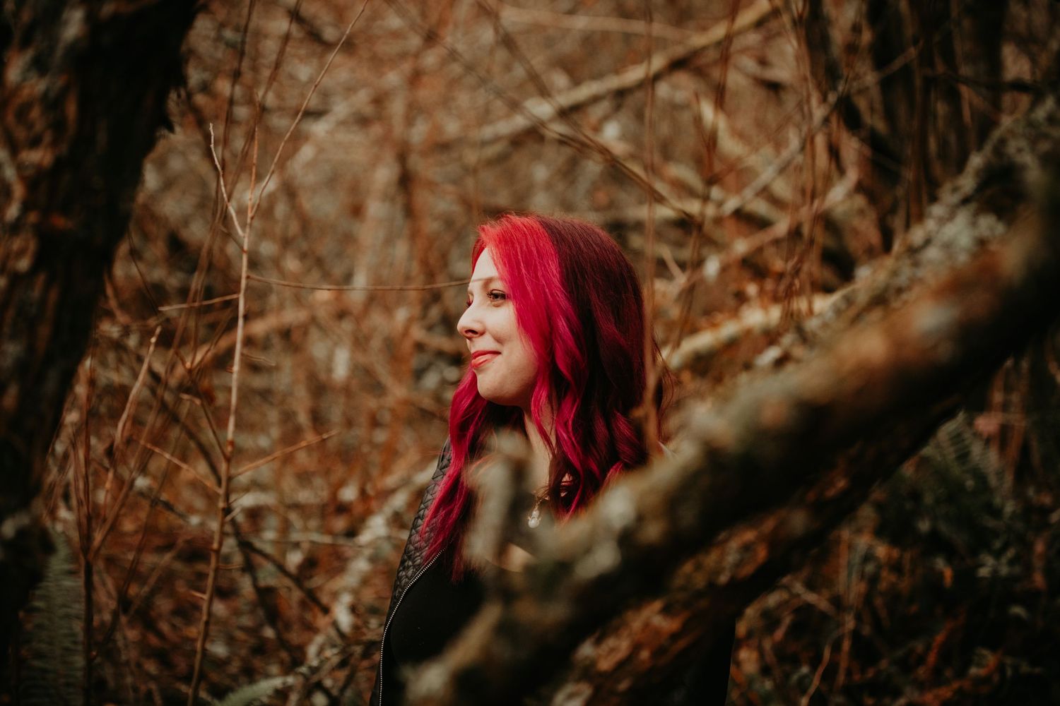 A person with vibrant red hair photographed through tree branches in an autumn forest setting.