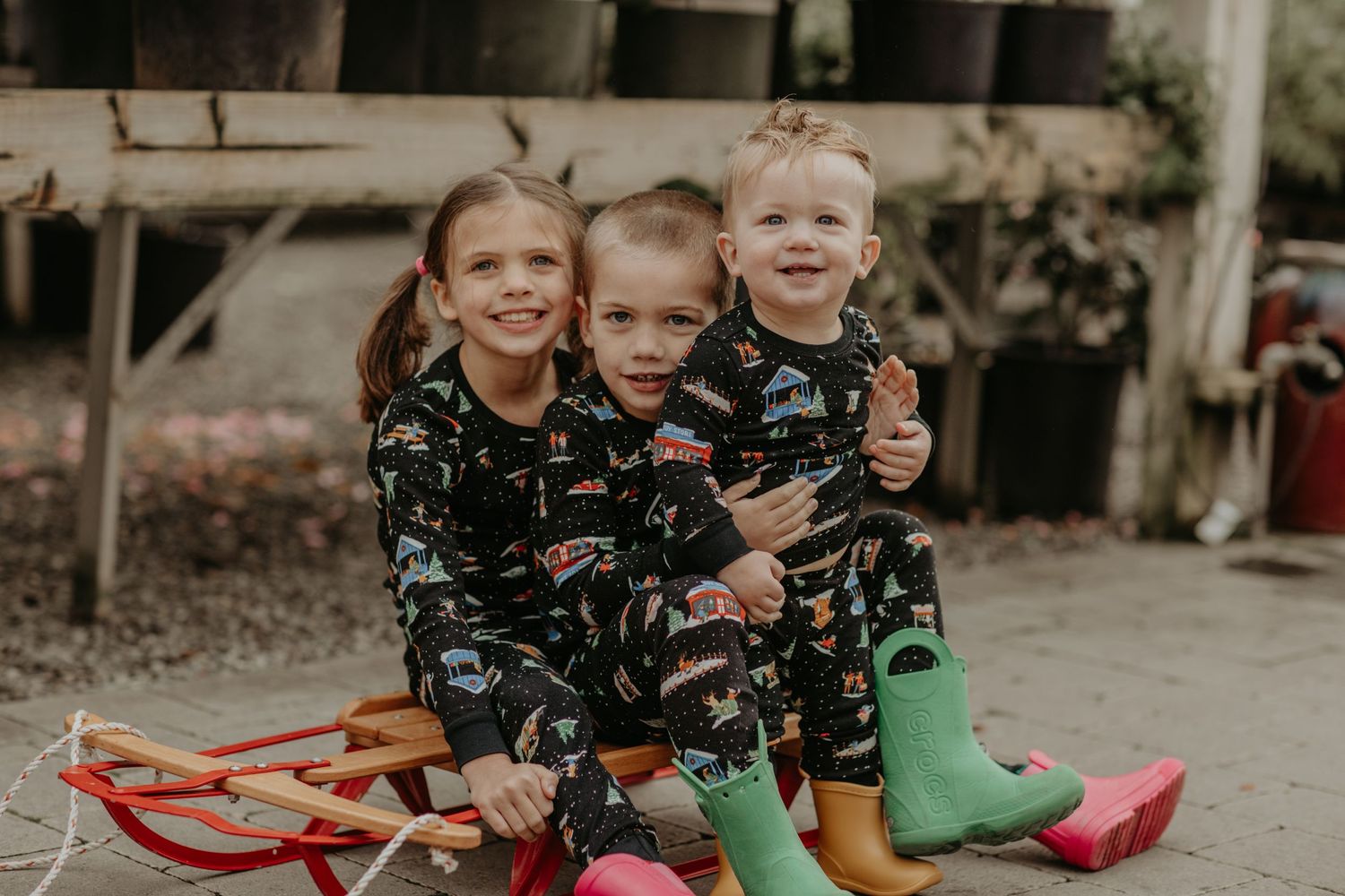 Children sit together on red sled wearing matching dinosaur pajamas and colorful rain boots for winter fun.