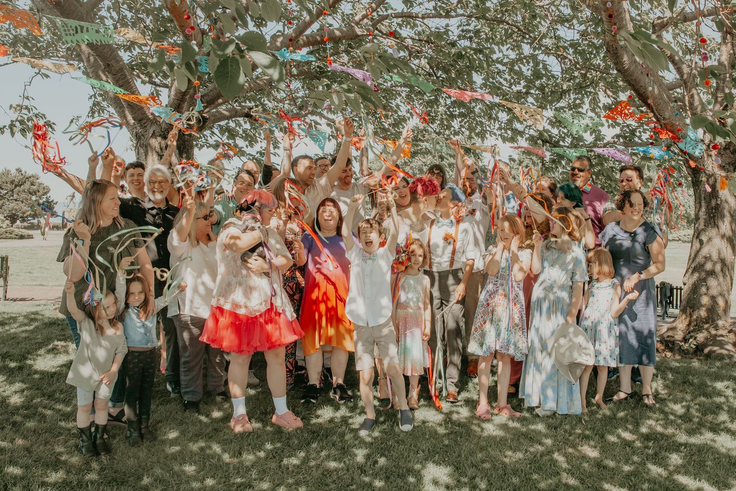 Large group of wedding guests celebrate outdoors under trees with colorful autumn foliage in the background.