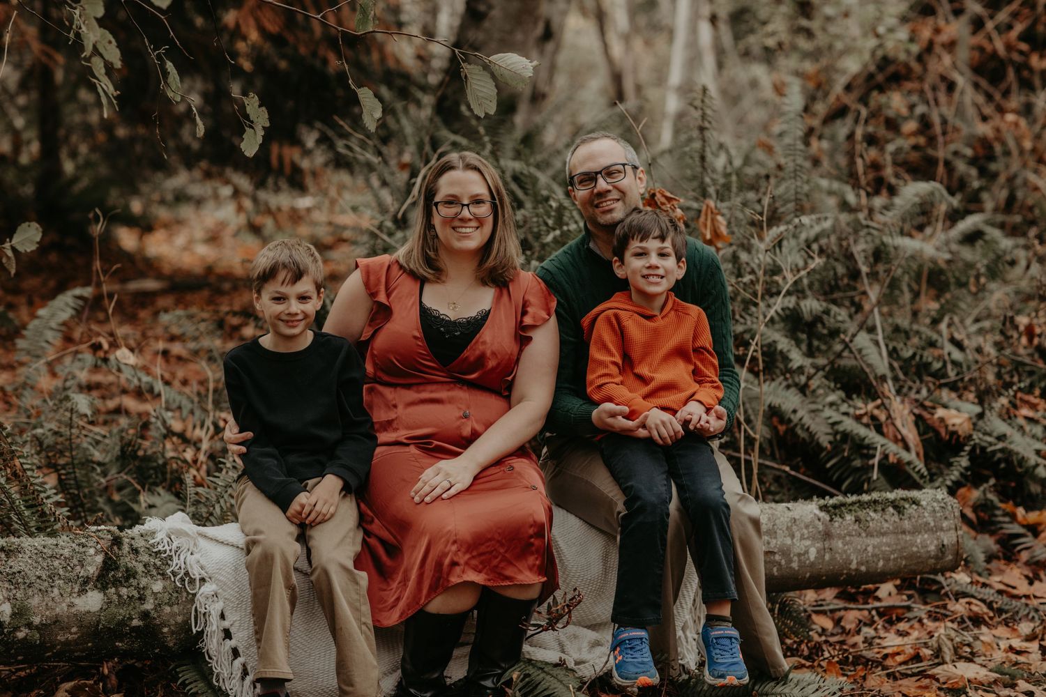 A family sits together on rocks in an autumn forest setting wearing coordinated casual outfits in warm fall colors.