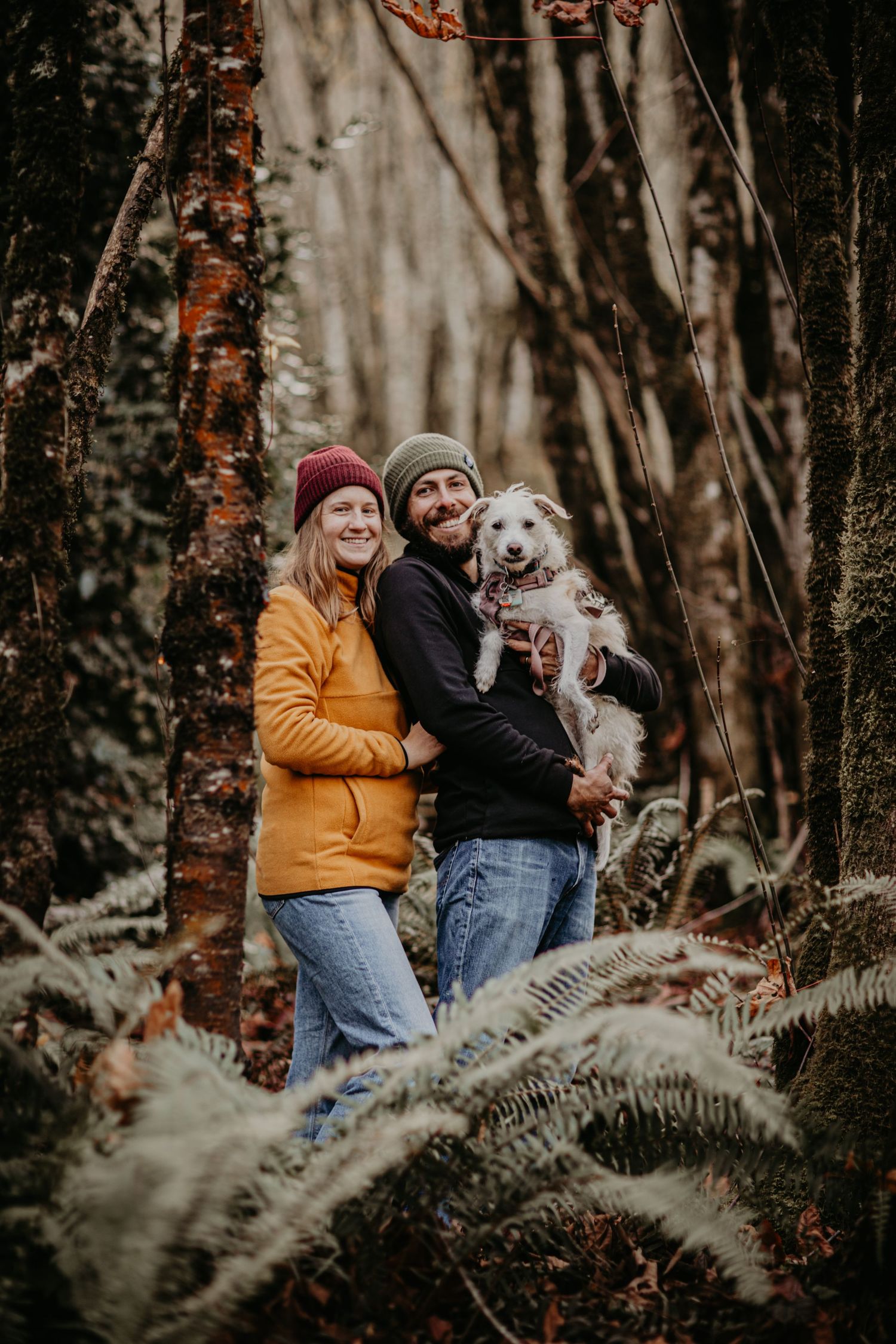 A couple wearing winter clothing stands in a forest with their white fluffy dog amid autumn foliage.