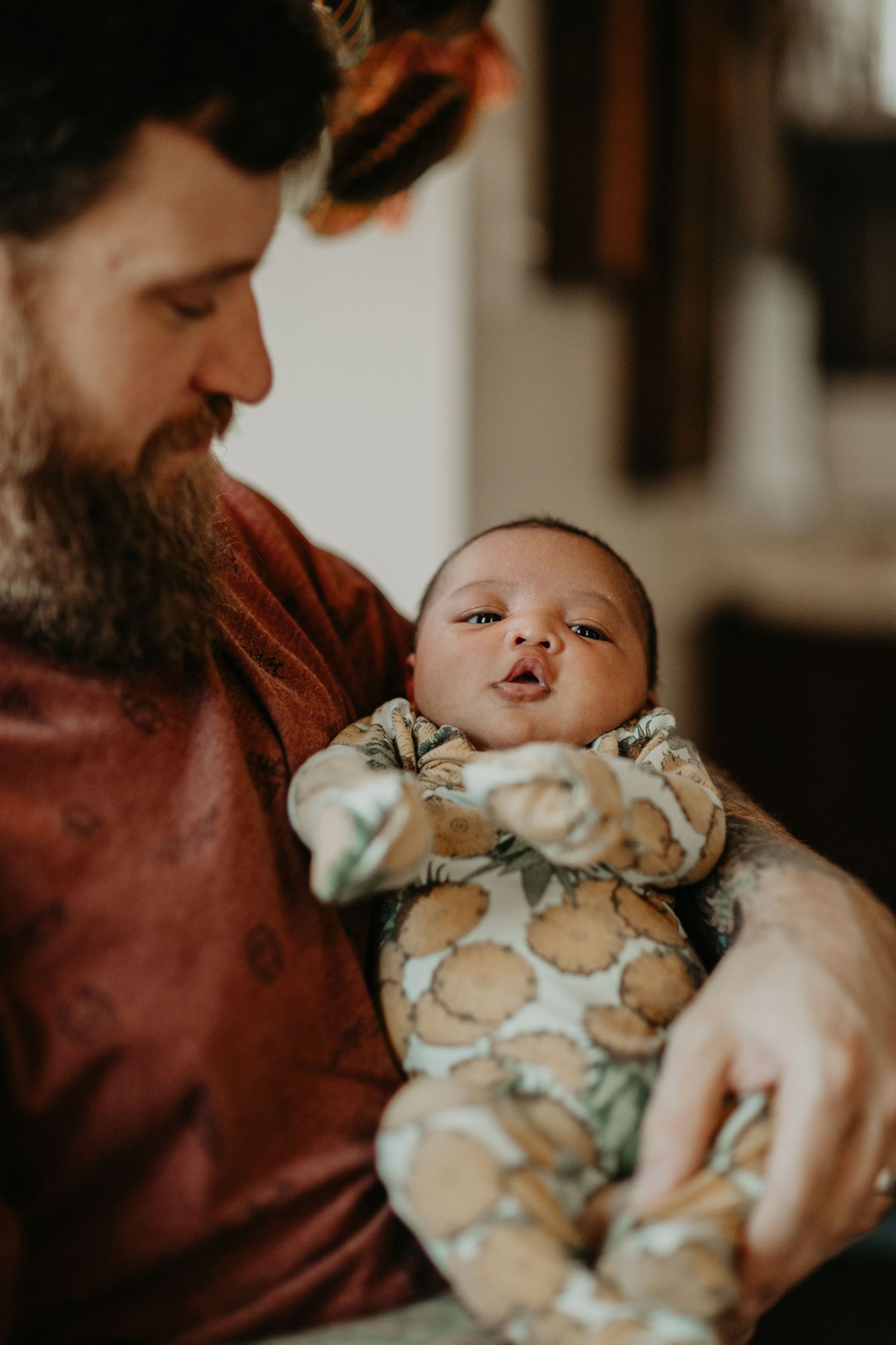 A bearded parent in a burgundy shirt tenderly holds an infant wearing a patterned onesie in a warm, intimate moment.