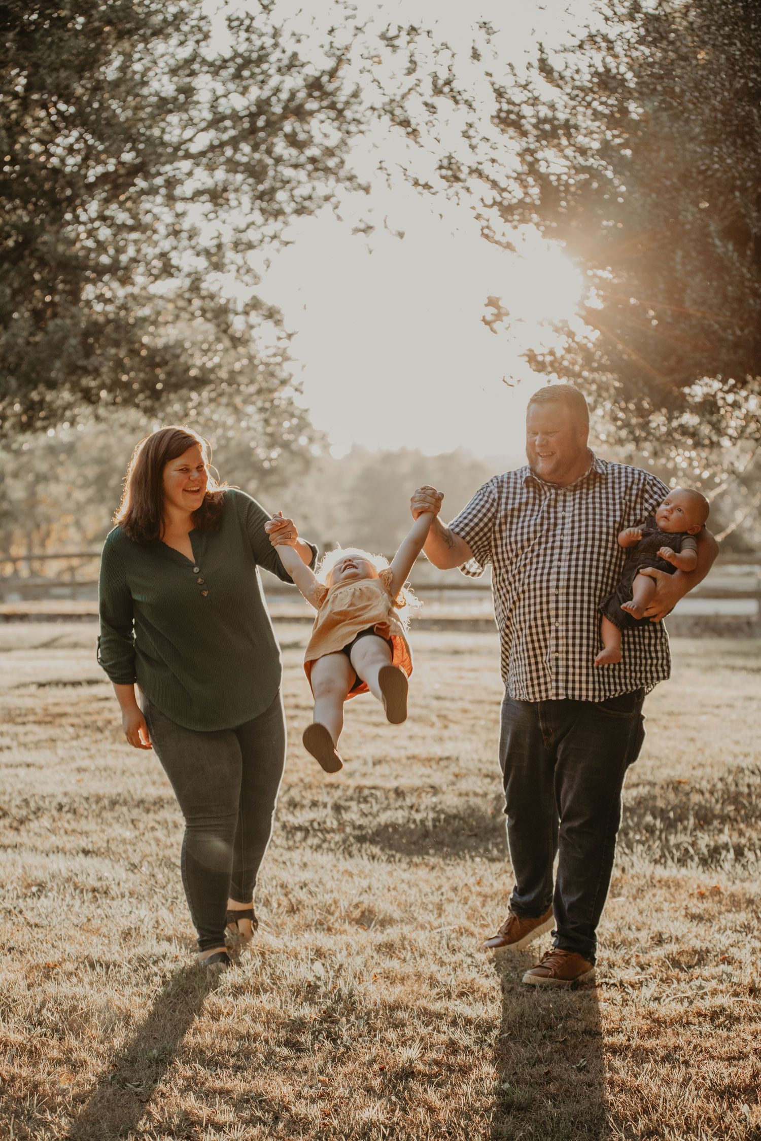 A family plays together in a sunlit field at golden hour, swinging a small child between them.