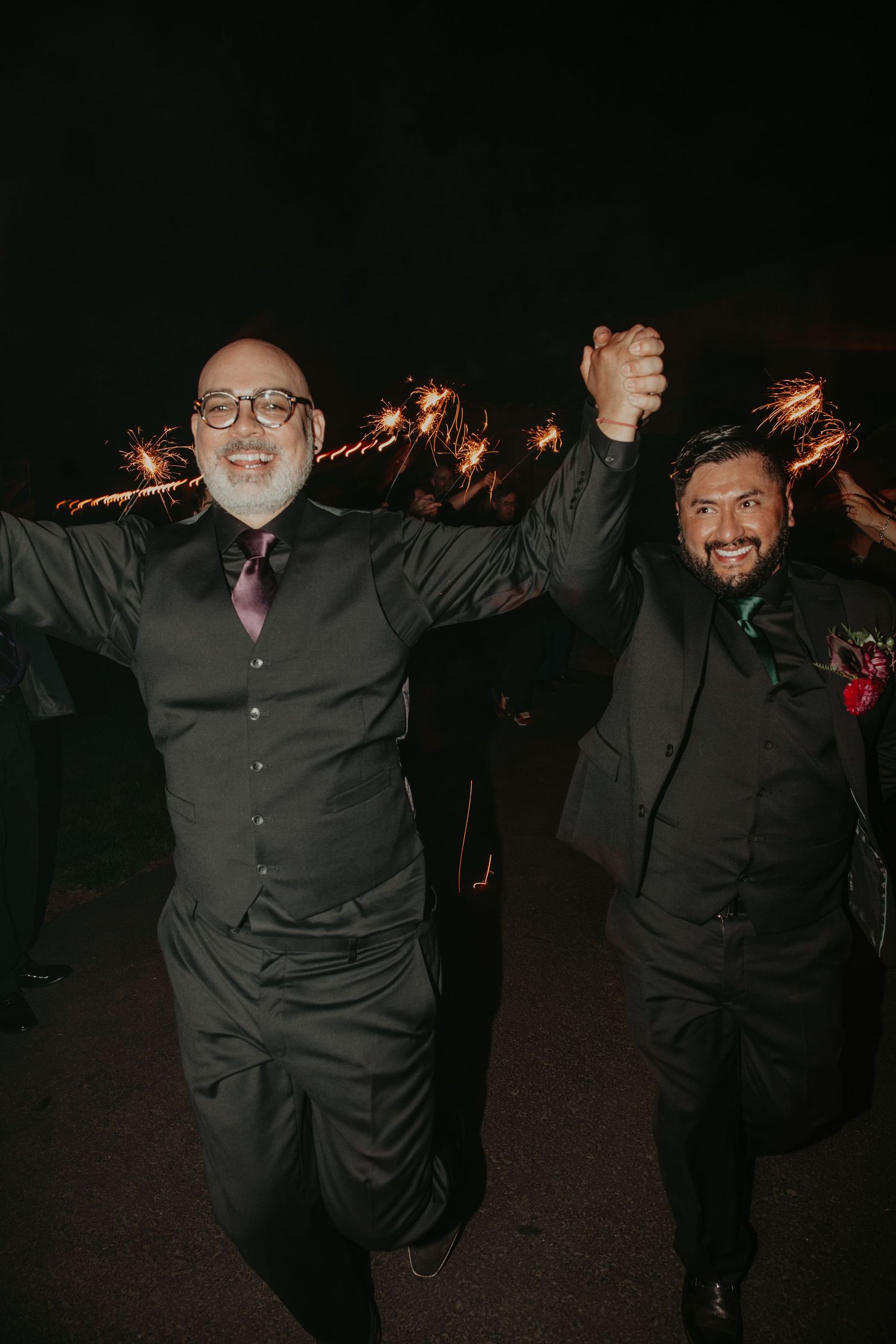Two men in dark suits dance joyfully at night with sparklers creating light trails in the background.