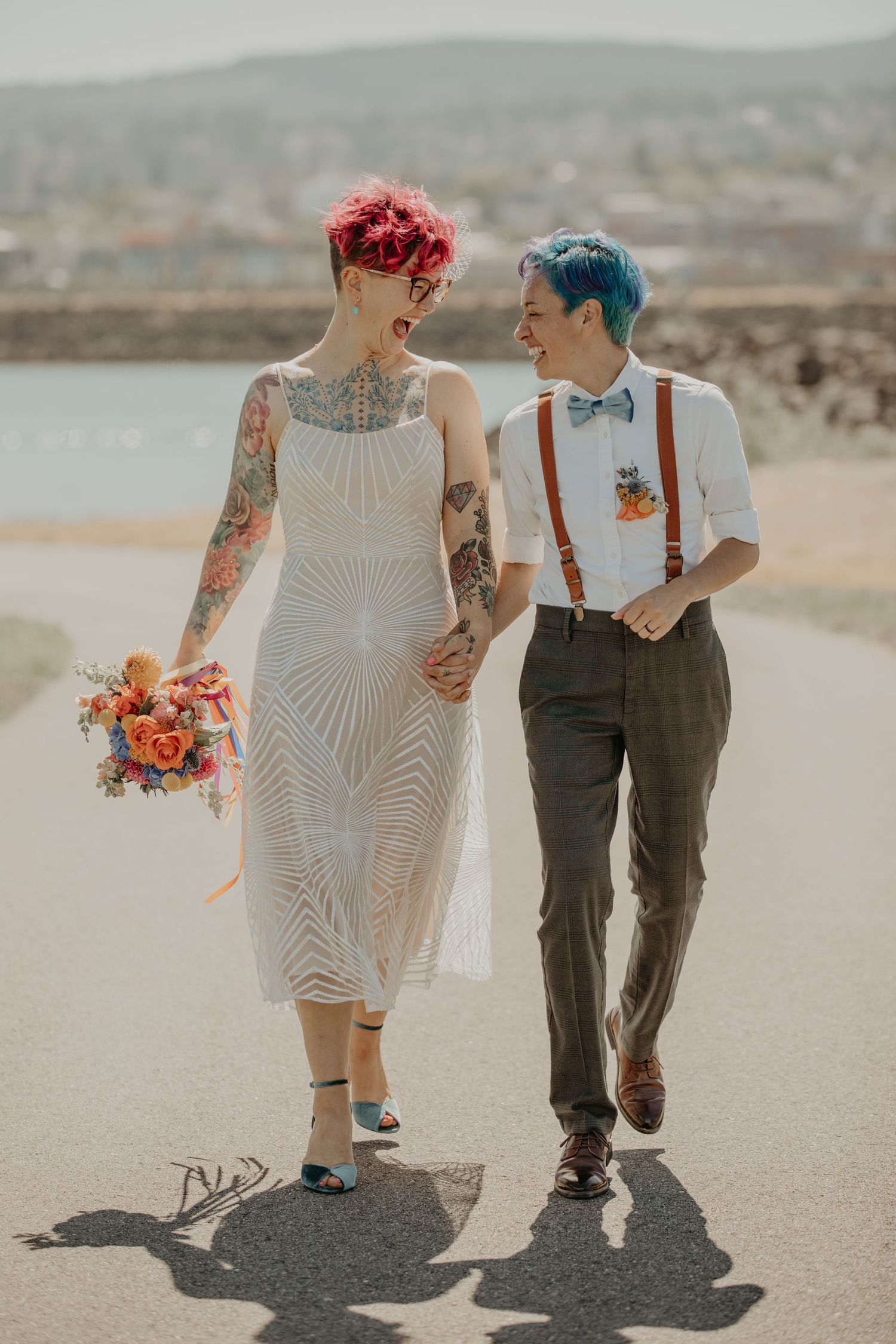 A couple in desert wedding attire walks together on a dusty path with mountains in the background.
