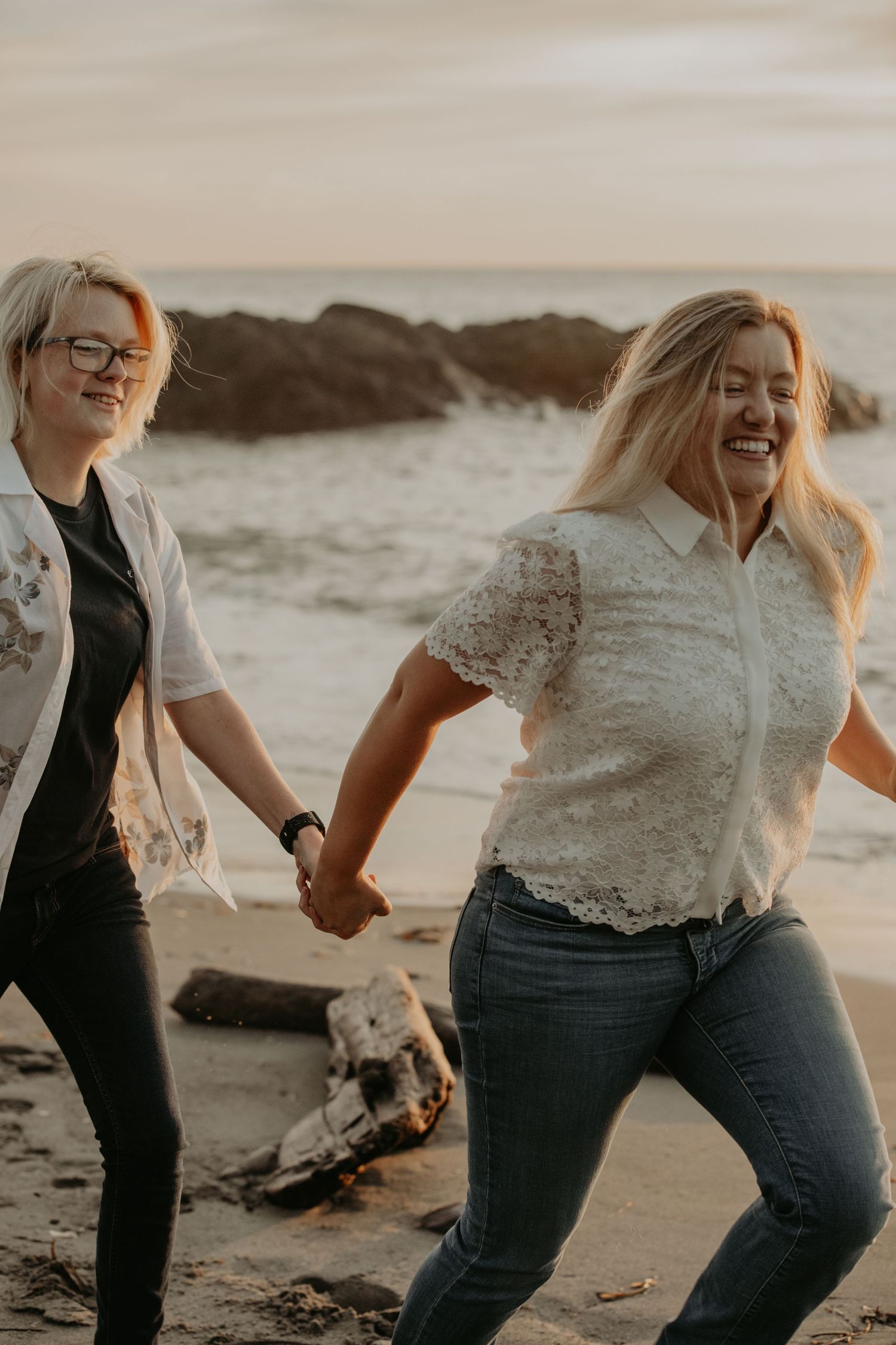Two people laughing and holding hands while walking on a beach at sunset, one wearing lace and the other casual attire.