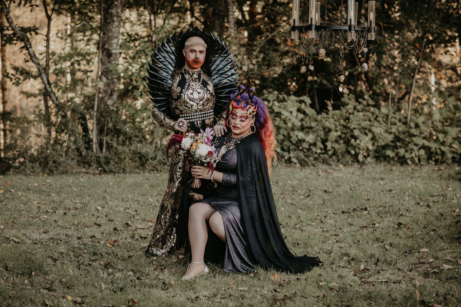 Two people in fantasy medieval costumes pose together in a grassy woodland setting for a themed photoshoot.