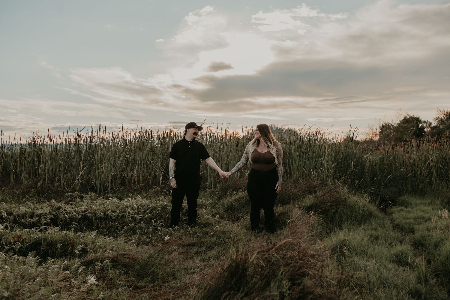 Two figures hold hands while standing on a scenic dirt path through tall grass at sunset.