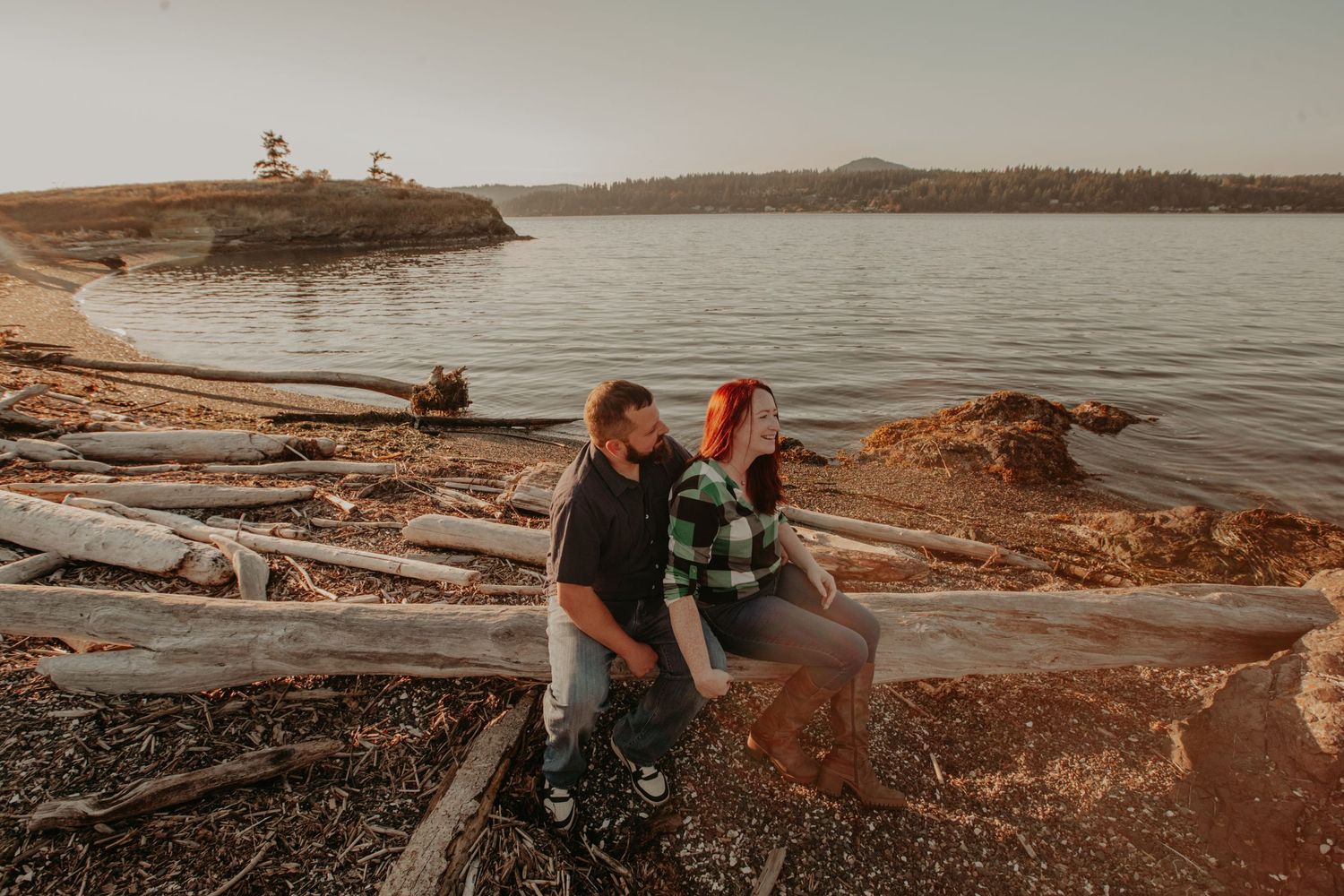 Two people sit together on driftwood along a rocky waterfront at sunset with mountains in the distance over calm waters.