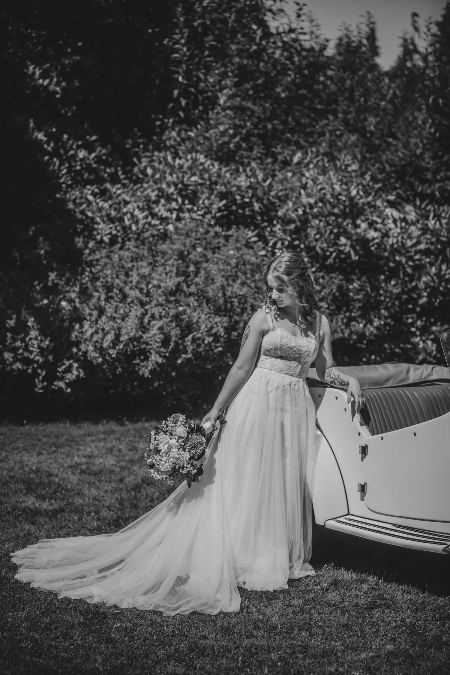 A bride in a flowing white gown poses beside a vintage car holding a bouquet.