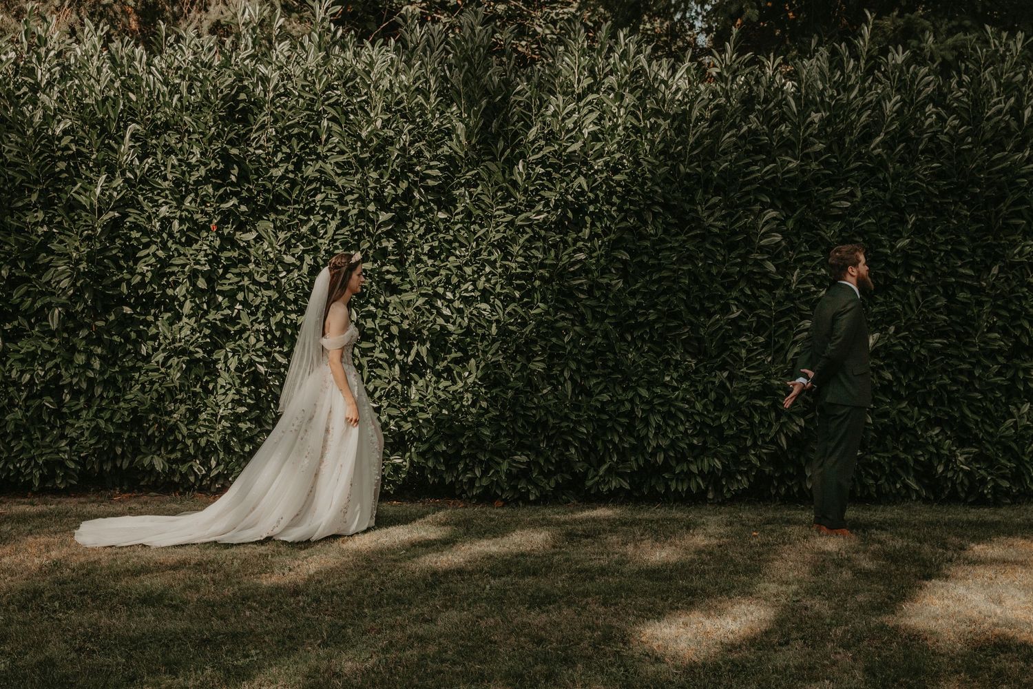 A bride in a white wedding dress walks along a lush garden path lined with tall green hedges.