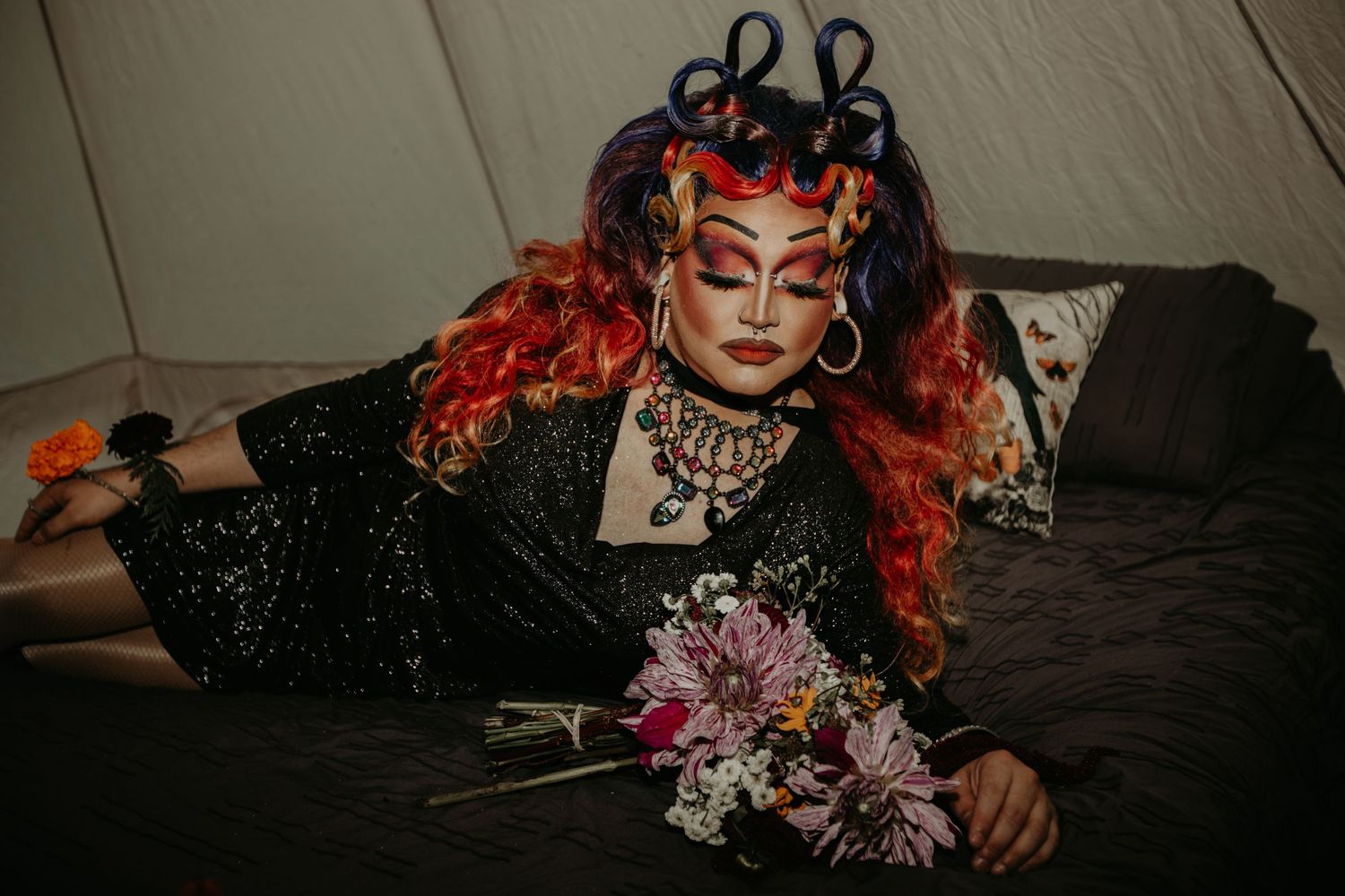 A person in gothic makeup and black dress poses with red hair and dried flowers on a dark surface.