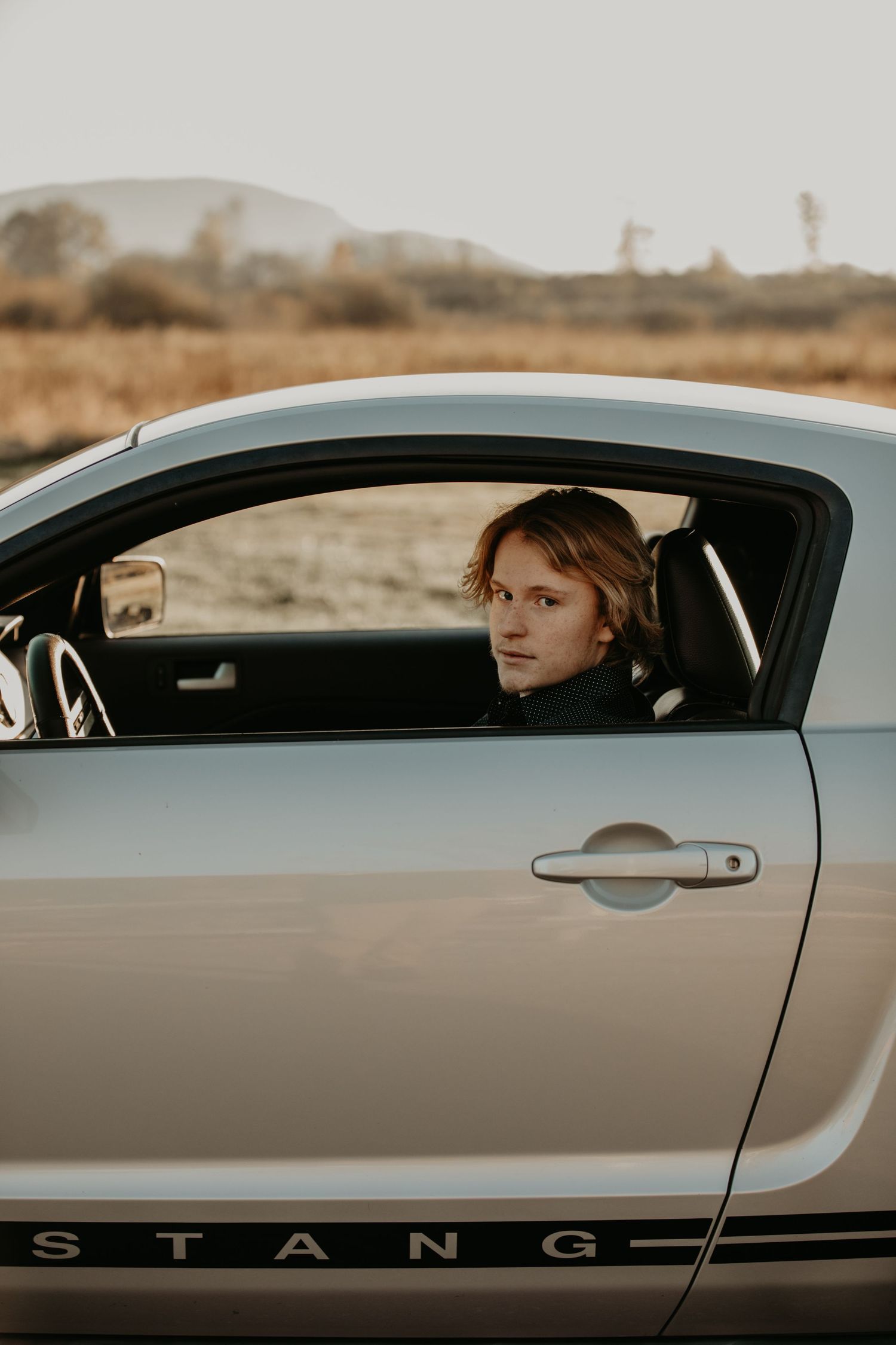 Side view of silver Mustang car with someone visible through driver's window in warm sunset lighting.