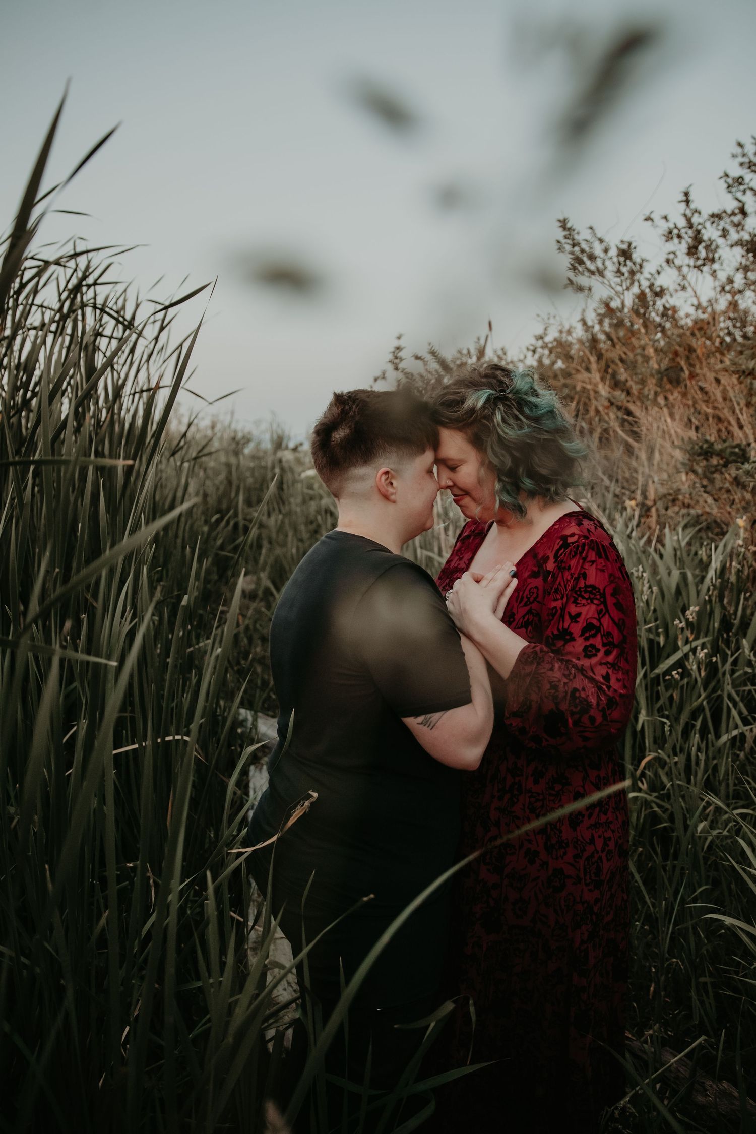 Romantic couple embraces in tall grass at sunset wearing red dress and black shirt in moody nature setting.