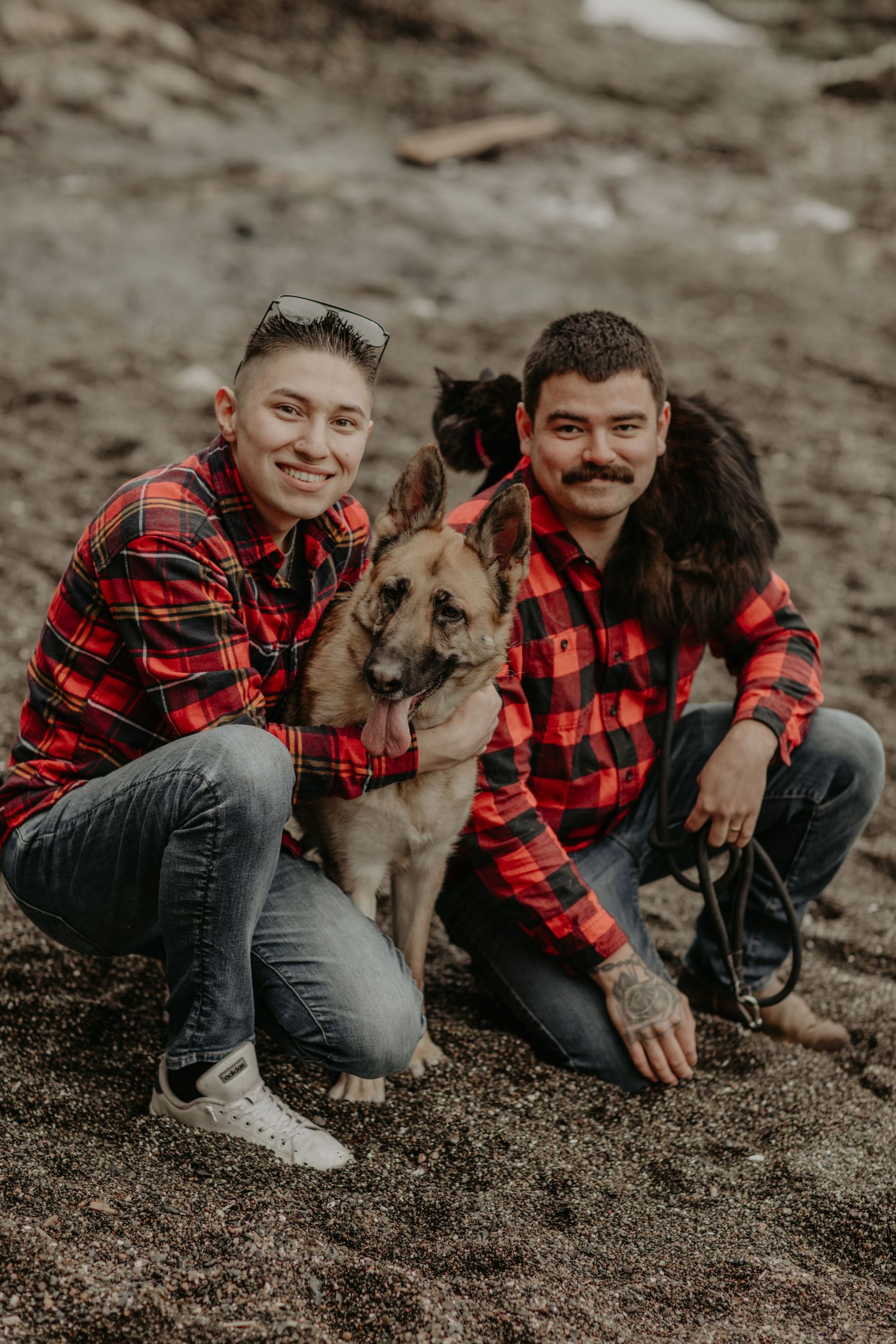 Two people in matching red buffalo plaid shirts pose outdoors with a German Shepherd dog on rocky terrain.