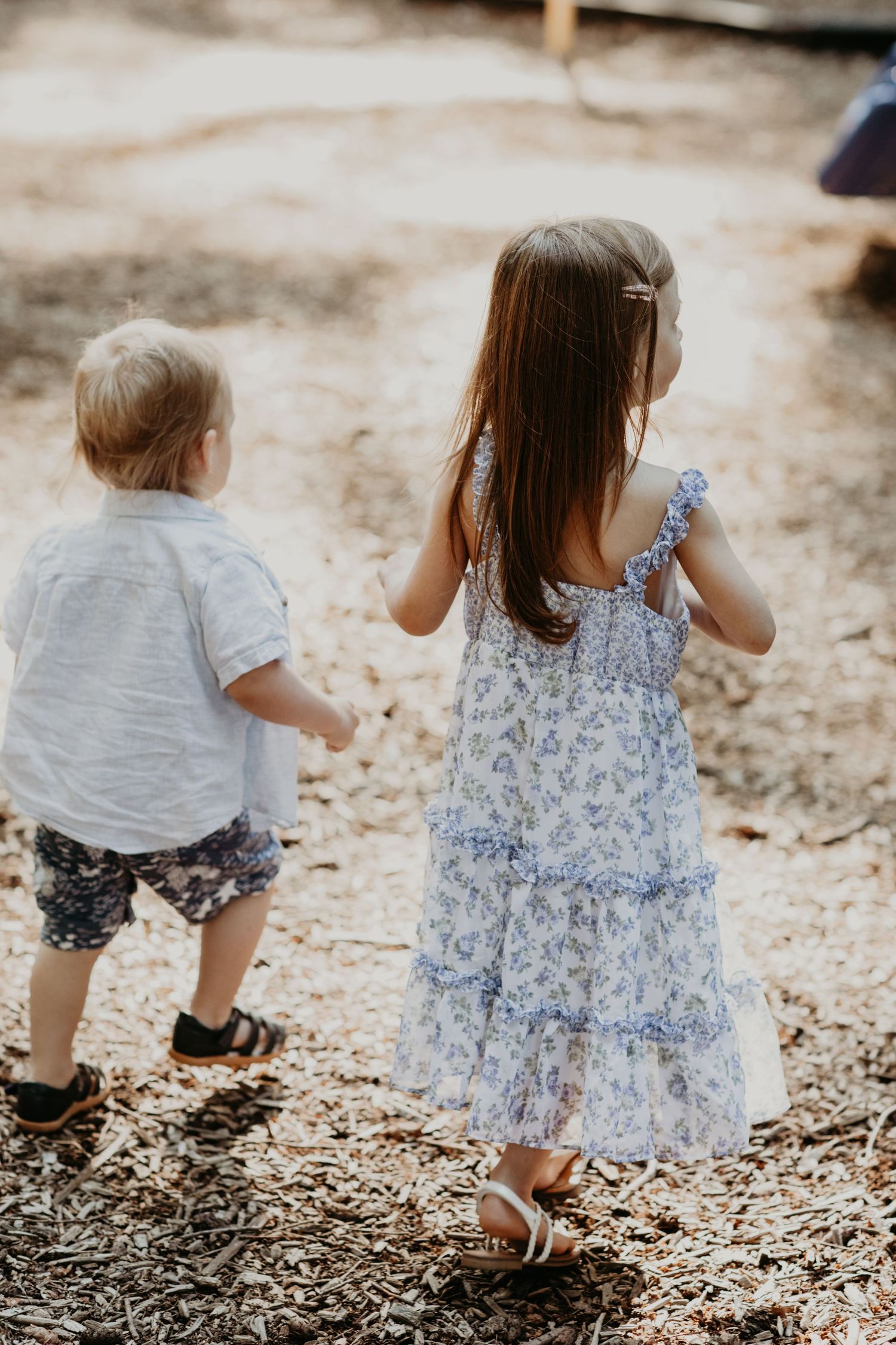 Two young children in summer outfits walk together on a dirt and mulch covered ground outdoors.